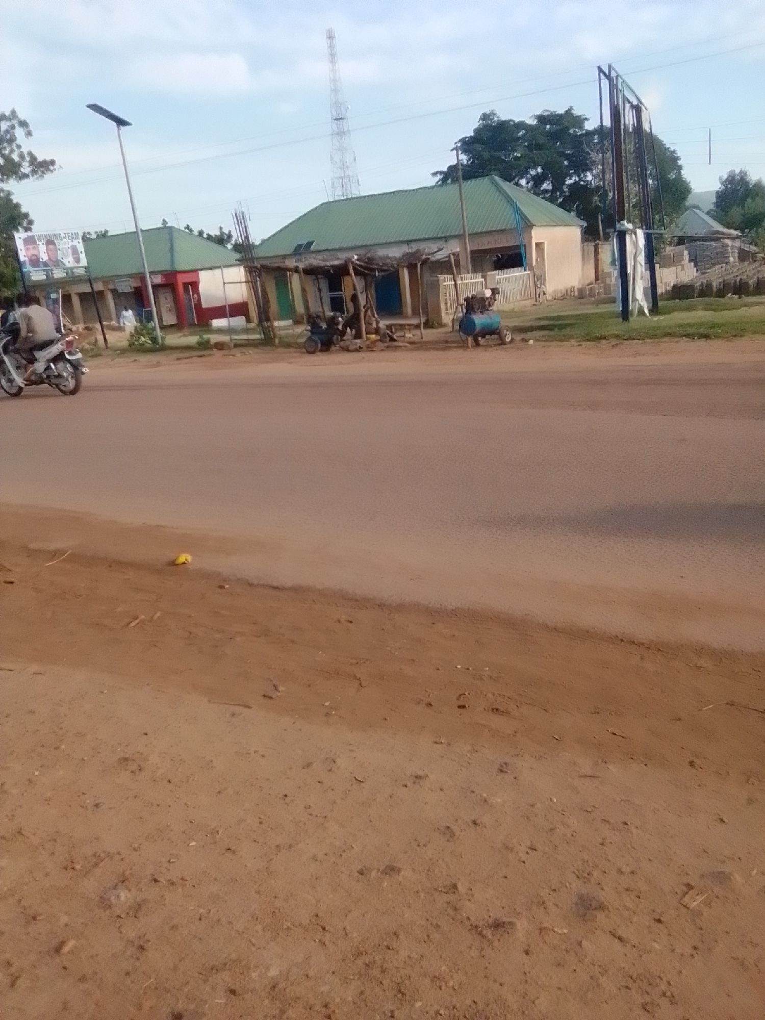 The image depicts a street scene in Galadima, Nigeria, with a paved road dominating the foreground and middle ground. On the left side of the road, a motorcycle with a rider is visible, heading away from the camera. Beyond the road, a row of buildings with green roofs lines the street. These buildings appear to be shops or small businesses, some with open fronts. A prominent billboard with images of three men and text, partially obscured, is attached to one of the buildings. To the right of the buildings, a tall telecommunications tower can be seen in the distance. Further to the right, an open area with stacked construction materials, possibly bricks, and a blue air compressor are present. The environment is dusty, with dry soil and scattered debris on the roadside. Trees and vegetation are visible behind the buildings, and the sky is overcast.