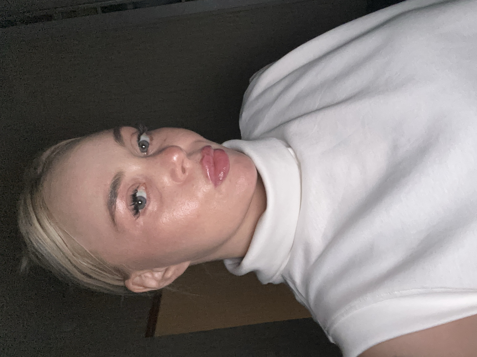A woman with blonde hair and light-colored eyes is looking towards the camera in a selfie. She has a light complexion and is wearing a white turtleneck top. Her lips are slightly parted and appear to be glossy. The background is dark and out of focus, with some indistinct shapes that could be furniture or a wall. There is no discernible information about the location in the city of Unknown, Unknown.