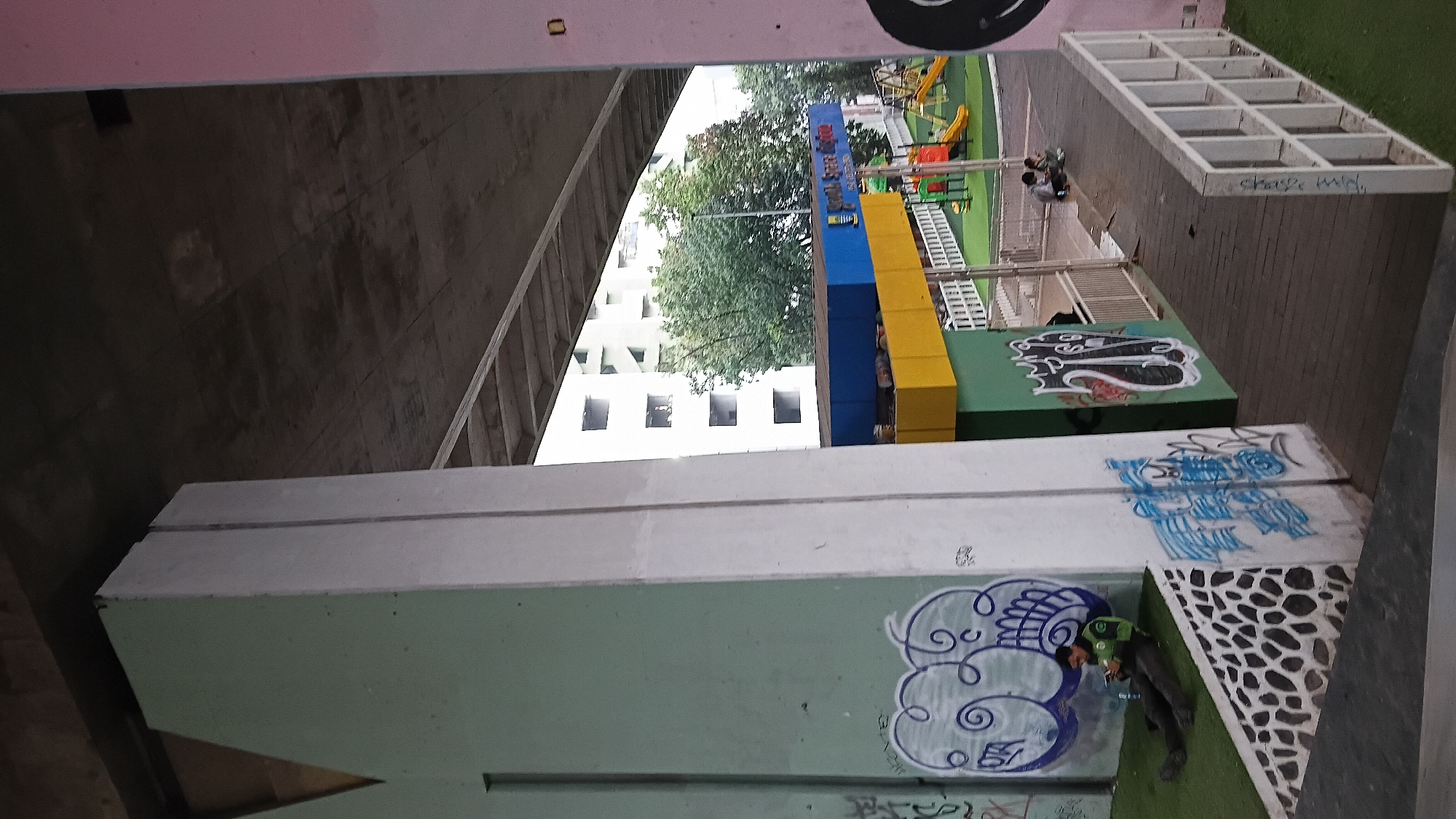 The image depicts an underpass area in Bandung City, Indonesia, with graffiti decorating concrete pillars and walls. To the left, a person in a green uniform is sitting on artificial grass, looking at their phone. In the background, a playground with slides and swings is visible, along with a building and trees. A wooden walkway with a white railing leads towards the playground. Another group of people is seated near the playground. The overall scene suggests an urban public space that has been adapted for recreational use.