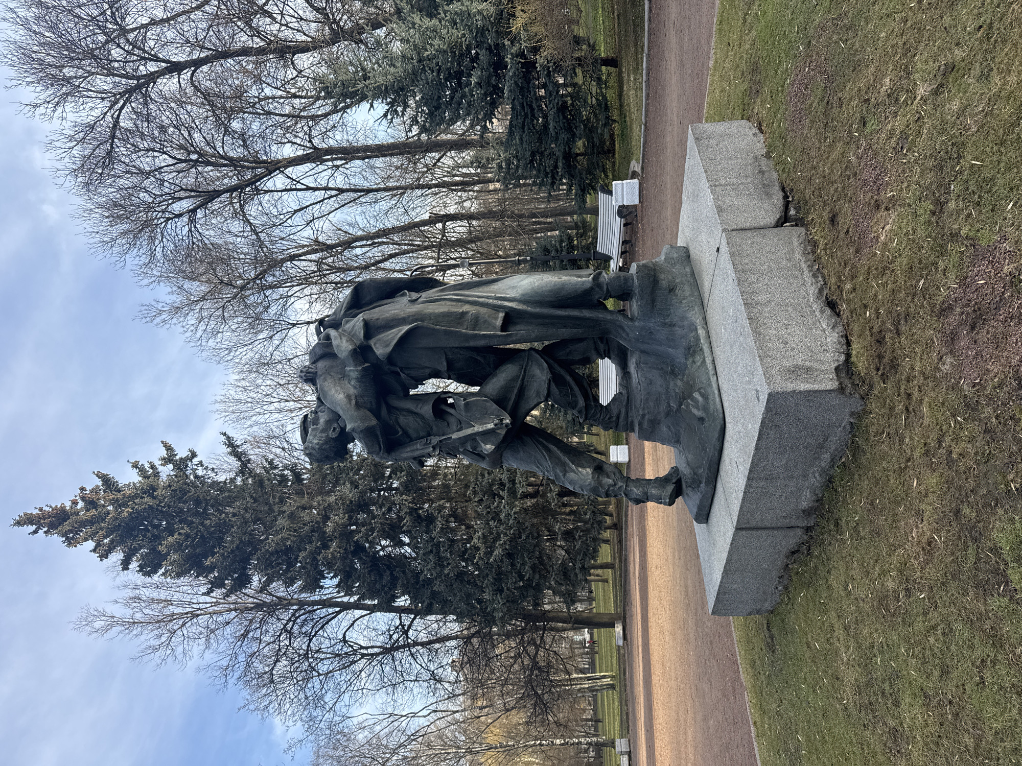 The image captures the "Grieving Mother" monument in Saint Petersburg, Russia. The monument depicts two soldiers, one carrying the other, in a somber, bronze-like color. It's mounted on a grey stone base, in a park setting. Bare trees stand tall in the background, with a few evergreen trees mixed in. A bench is also seen in the background. There's a patch of grass to the right of the statue, and the sky appears overcast. A dirt path is seen to the right of the frame.