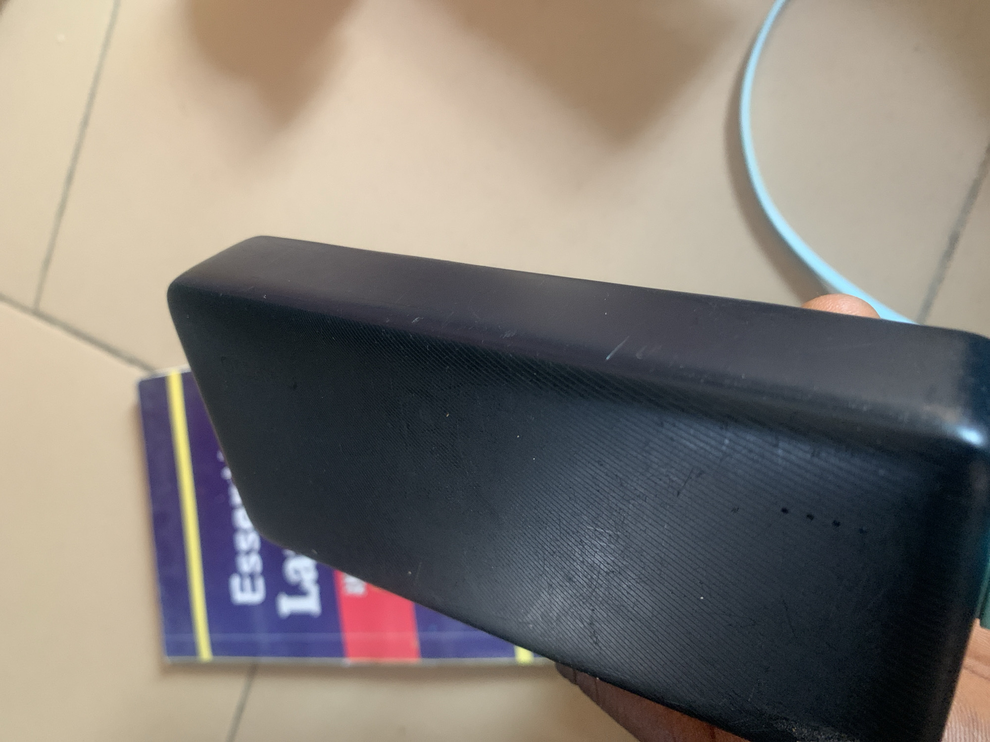 In Jauro Yinu, Nigeria, a black power bank is held in a person's hand, partially obscuring a book with the title "Essentials of Law". The power bank has a textured surface with visible scuffs and scratches. Four small dots, likely indicator lights, are present on its surface. A light blue cable is visible to the right of the power bank. The background appears to be a light-colored floor.