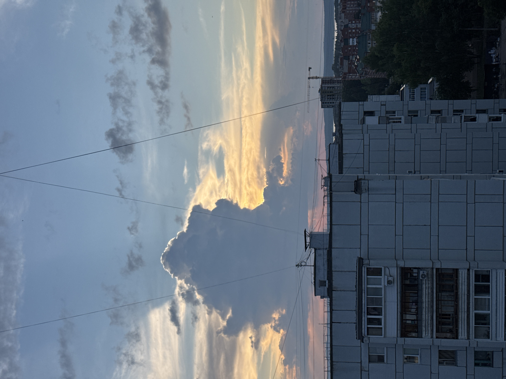 The image depicts a cloudy sky at dusk over the city of Chaikovsky, Russia. In the foreground, the tops of several apartment buildings are visible. The closest building is a multi-story structure with balconies, its facade made of grey concrete panels. Above the buildings, thin wires crisscross the sky. A large, dramatic cloud formation dominates the upper half of the image, illuminated from behind by the setting sun, which casts a warm, orange glow. In the distance, other buildings and trees can be seen, and beyond them, a body of water. A construction crane is visible in the mid-distance. The scene appears to be a typical urban landscape under a dramatic sky.