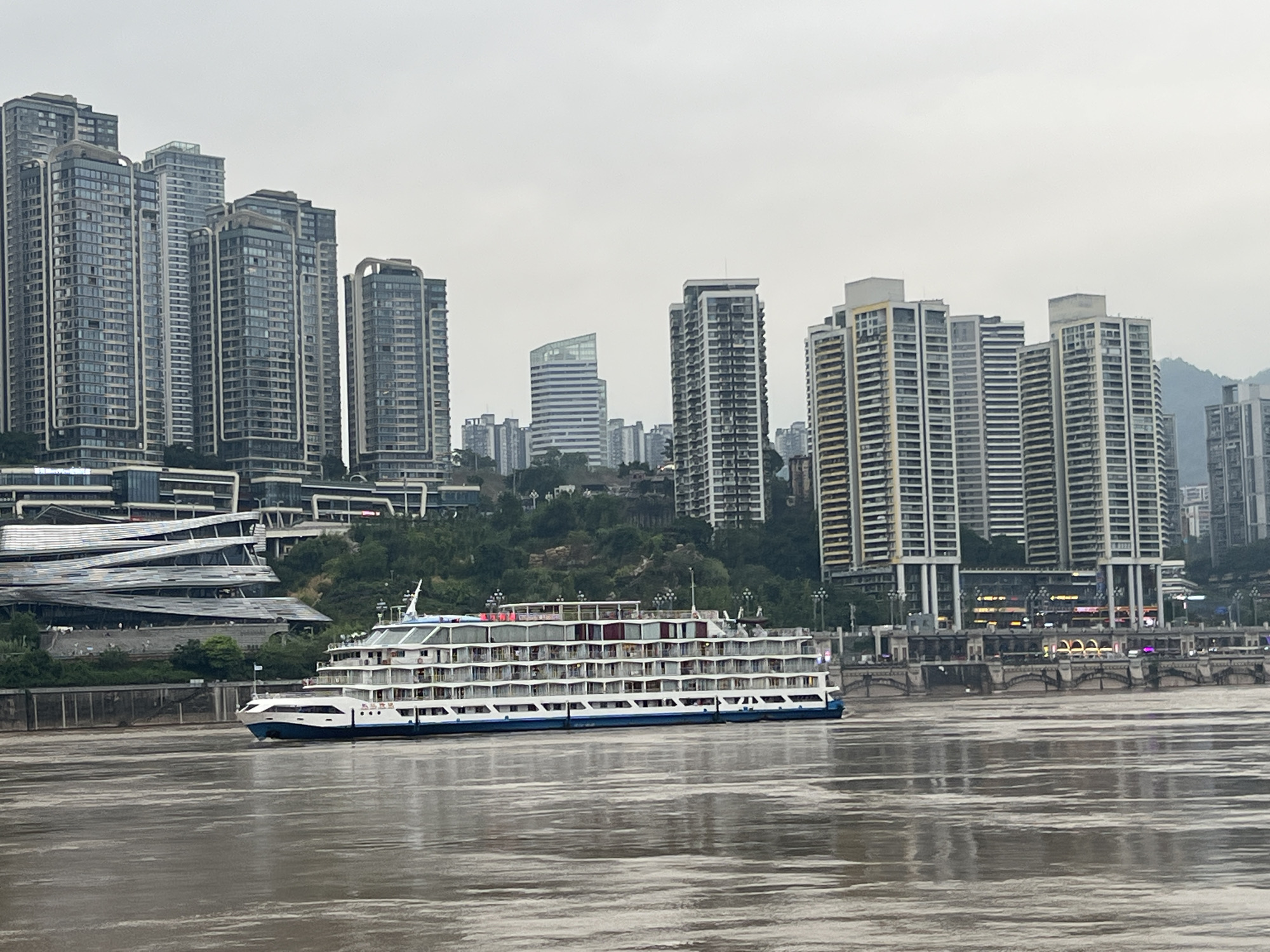 In the Yuzhong District of China, a large, multi-decked white cruise ship with blue trim is navigating a wide, murky river. The vessel is positioned in the foreground, moving from left to right. Behind the ship, a densely packed urban skyline rises, dominated by numerous tall, modern apartment buildings and office towers constructed with glass and concrete. The architecture varies, with some buildings featuring sleek, contemporary designs and others appearing more traditional. Several bridges are visible in the background, spanning the river and connecting different parts of the city. Lush green trees and vegetation are interspersed between the buildings and along the riverbanks, indicating a blend of urban development and natural scenery. The sky is overcast and a pale gray, suggesting a cloudy or hazy day. The water of the river reflects the muted tones of the sky and the surrounding structures. The scene captures a bustling city on a waterway, with the cruise ship as a central element.