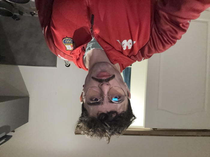 A man with glasses and a mustache wears a red Kappa jacket with an Italian Olympic emblem on the chest. He looks directly at the camera with a neutral expression. His hair is curly. The background is a plain white wall with a glimpse of a kitchen counter on the left. The image appears to be inverted.