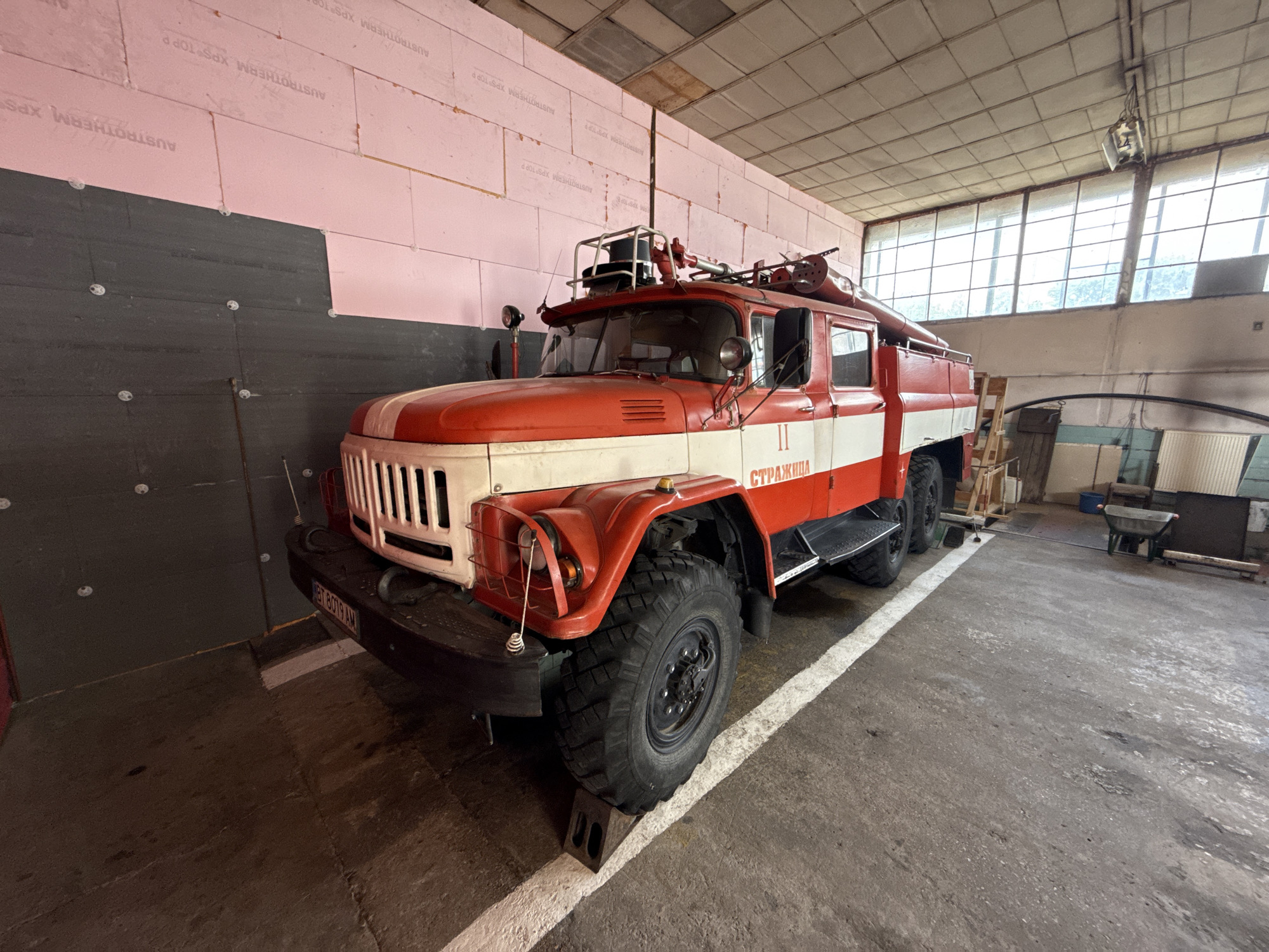 The image shows a vintage red and white fire truck parked inside a large, industrial-style building. The truck has the word "СТРАЖИЦА" (Strazhitsa in Bulgarian) written on its side, indicating it likely belongs to the city of Strazhitsa in Bulgaria. The vehicle also displays a license plate reading "BT 8019 AM".
The building's walls are lined with pink insulation panels and dark gray material, possibly soundproofing or additional insulation. The floor is concrete, and a white line is painted on the floor next to the tire of the vehicle. In the background, there's a window and some equipment and debris. The lighting is mostly provided by sunlight coming through the windows.