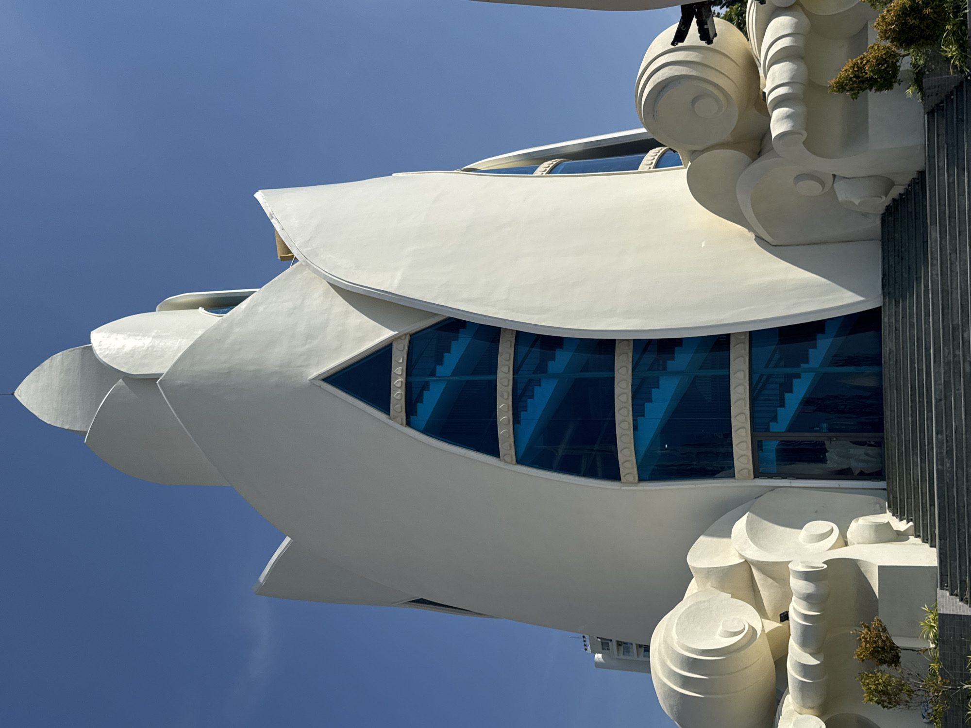 The image displays the front facade of the Long Son Pagoda in Nha Trang, Vietnam. The pagoda features a distinctive white, lotus-shaped architectural design with large, blue glass windows revealing a staircase. The exterior is adorned with intricate white sculptural elements resembling swirling clouds or waves on either side of the entrance. In the foreground, stone steps lead up to the pagoda's main entrance, flanked by small, green trees and a gray tiled walkway. The sky above is a clear, bright blue. The scene is captured under daylight, with strong shadows indicating the sun's position.