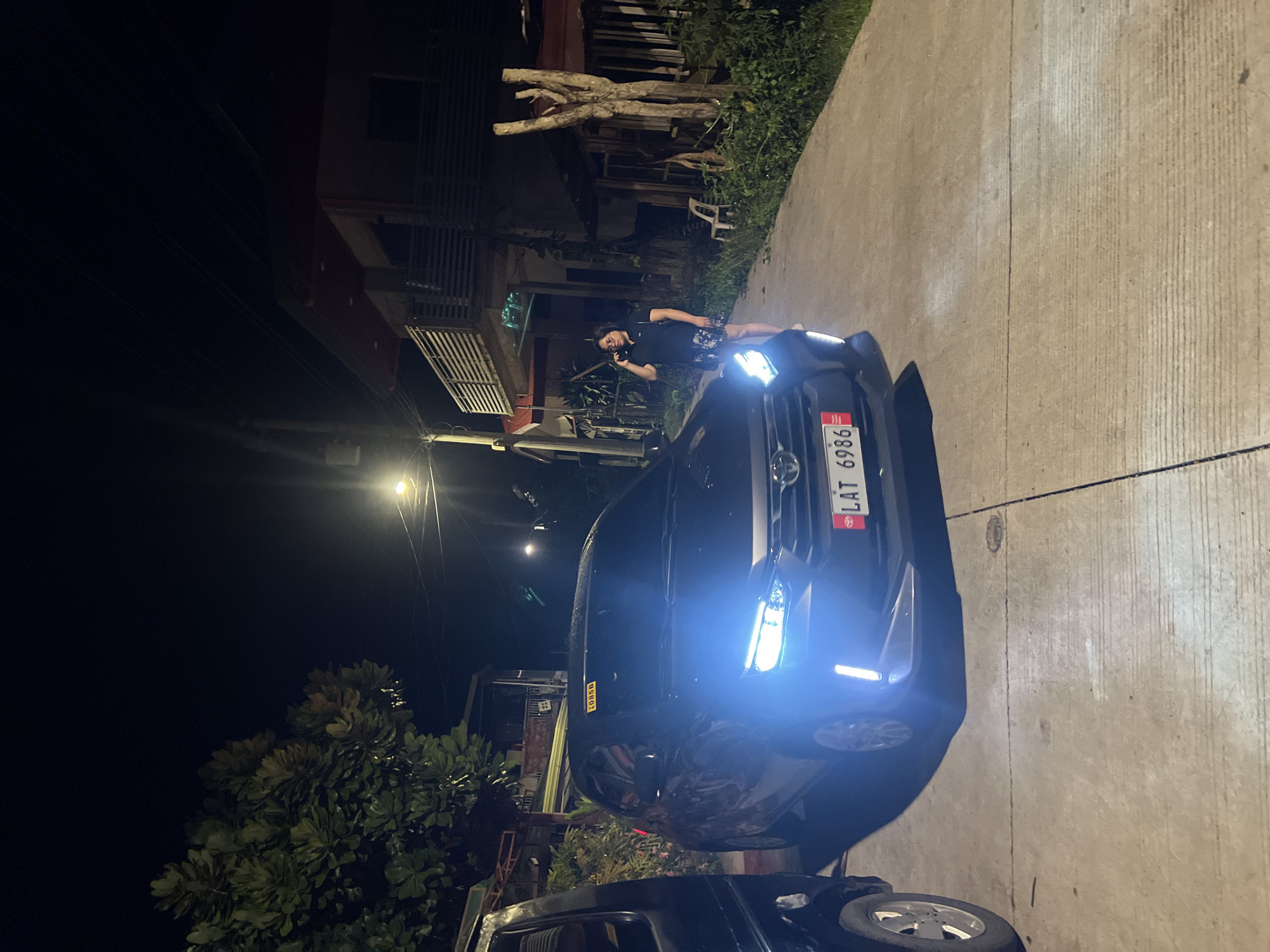 A dark, outdoor night scene in Davao City, Philippines, presents a black car with illuminated headlights parked on a concrete street. A man stands by the driver's side door, holding a cellphone to his ear. A Philippine license plate, "LAT 6986," is visible on the front of the vehicle. Buildings and vegetation are visible in the background, under a street light.