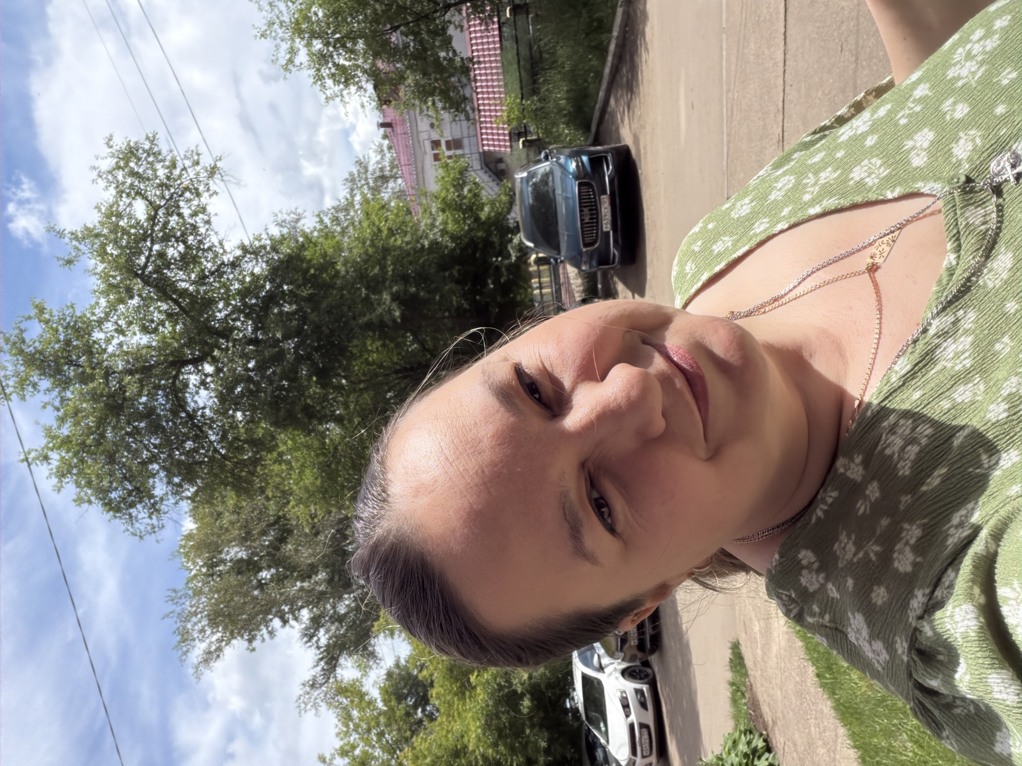 A woman is taking a selfie outdoors on a sunny day. She is wearing a green dress with a floral pattern and two necklaces. In the background, there are trees, parked cars, and a building with a pink roof. The setting appears to be a residential street in the city of Chaikovsky, Russia.
