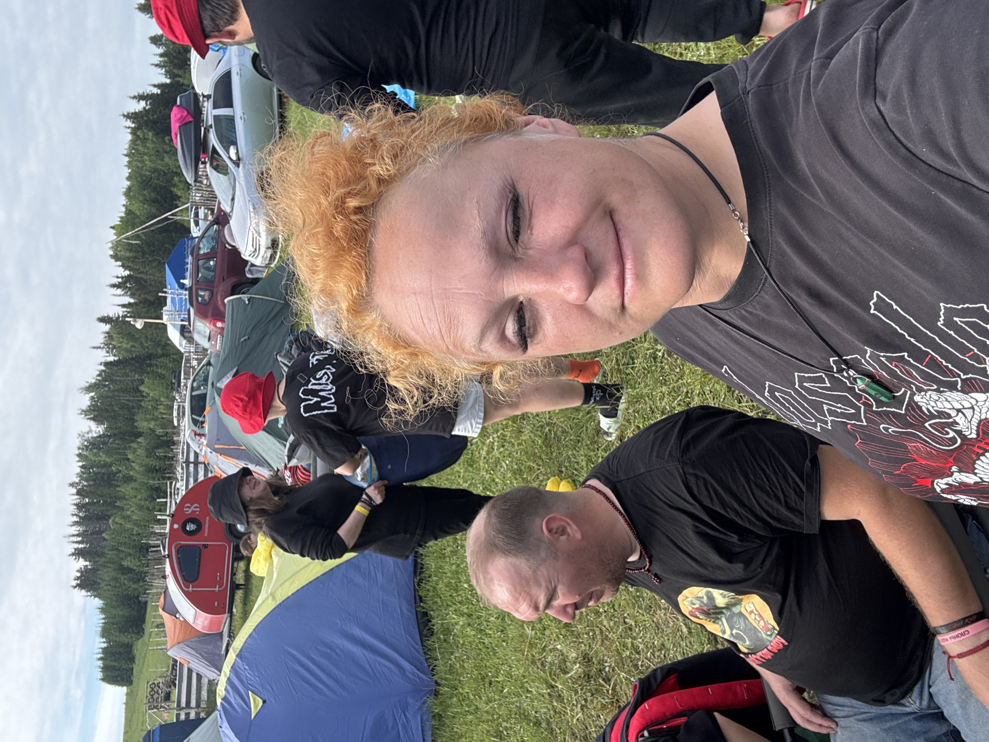 The image depicts an outdoor gathering in a grassy field in Большая Кивара, Russia, under an overcast sky, with a dense coniferous forest forming the distant background. In the foreground, a woman with curly red-orange hair, wearing a black t-shirt with a white graphic and a thin black necklace, faces the camera with a slight smile. To her left, a man with a receding hairline and a beard, wearing a black t-shirt with a yellow and red graphic, blue jeans, a pink wristband, and a dark red beaded necklace, is seated and looking towards the right. A red and black bag is positioned next to him.
In the mid-ground, multiple individuals are visible. A woman wearing a black bucket hat, black clothing, and a yellow wristband, holding a light-colored pouch, stands facing right, partially obscured by a large blue tent with yellow trim. Adjacent to her, another person, seen from the back, wears a red bucket hat, a black t-shirt with "MISFITS" written in white, white shorts, orange socks, and black and white sneakers. To the far right, partially obscured by the foreground woman's hair, a man in a red cap, black t-shirt, and black pants is visible from behind, wearing red flip-flops.
The field is populated with various camping tents, including orange, grey, and green structures. A prominent red teardrop-shaped camper trailer with a black "8" logo and "PANORAMA" text is centrally located. Numerous passenger vehicles, including a silver sedan with a black rooftop cargo box and a maroon SUV, are parked in the background. A wooden fence runs horizontally across the mid-distance, and a tall, thin pole extends upwards towards the sky in the far background.