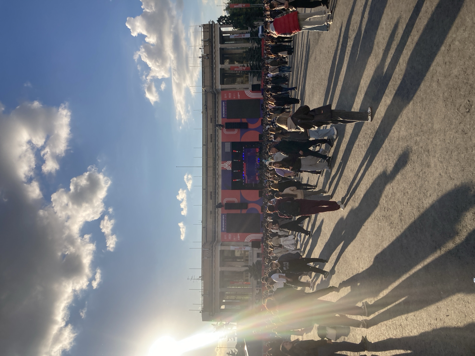 The image shows a crowd of people gathered in front of a building in Moscow, Russia. The building appears to be a public venue, possibly for sports or entertainment, based on the large screen and signage. The event appears to be a celebration or gathering, as people are standing and observing the event. The lighting suggests it is either early morning or late afternoon. The presence of long shadows indicates a low sun angle. The weather is sunny with scattered clouds.