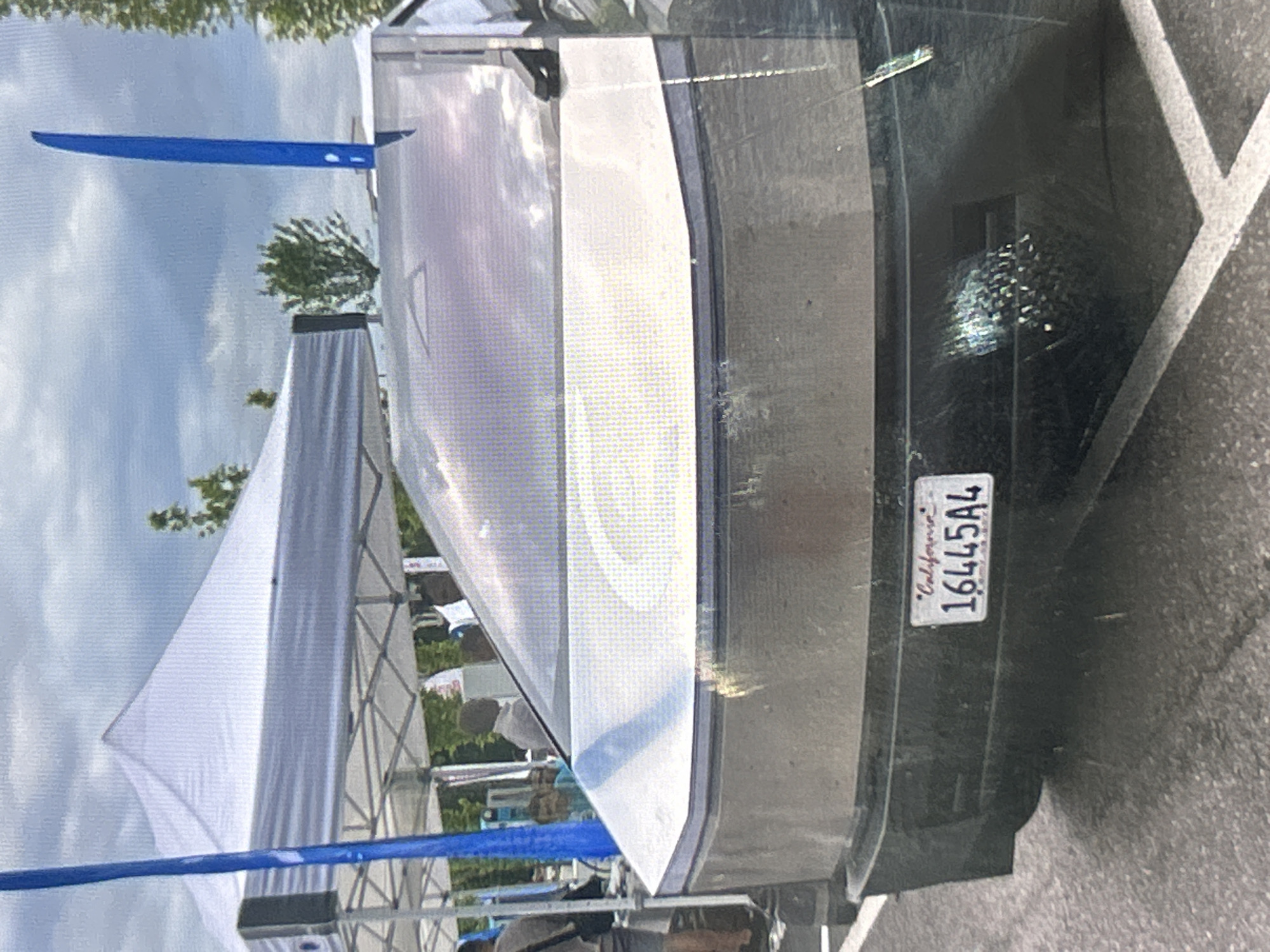 An outdoor scene under a cloudy sky shows the rear of a modern, angular, silver-colored vehicle. The vehicle has a large, tinted rear window that reflects the sky and surrounding environment. Below the window is a horizontal band of lighter silver, followed by a darker, matte black bumper area. A California license plate with the number "16445A4" is affixed to the bumper. To the left of the vehicle, a blue pole rises vertically, and behind it, a white tent structure is visible. Trees and a few individuals, mostly out of focus, can be seen in the background. The ground is paved with asphalt.