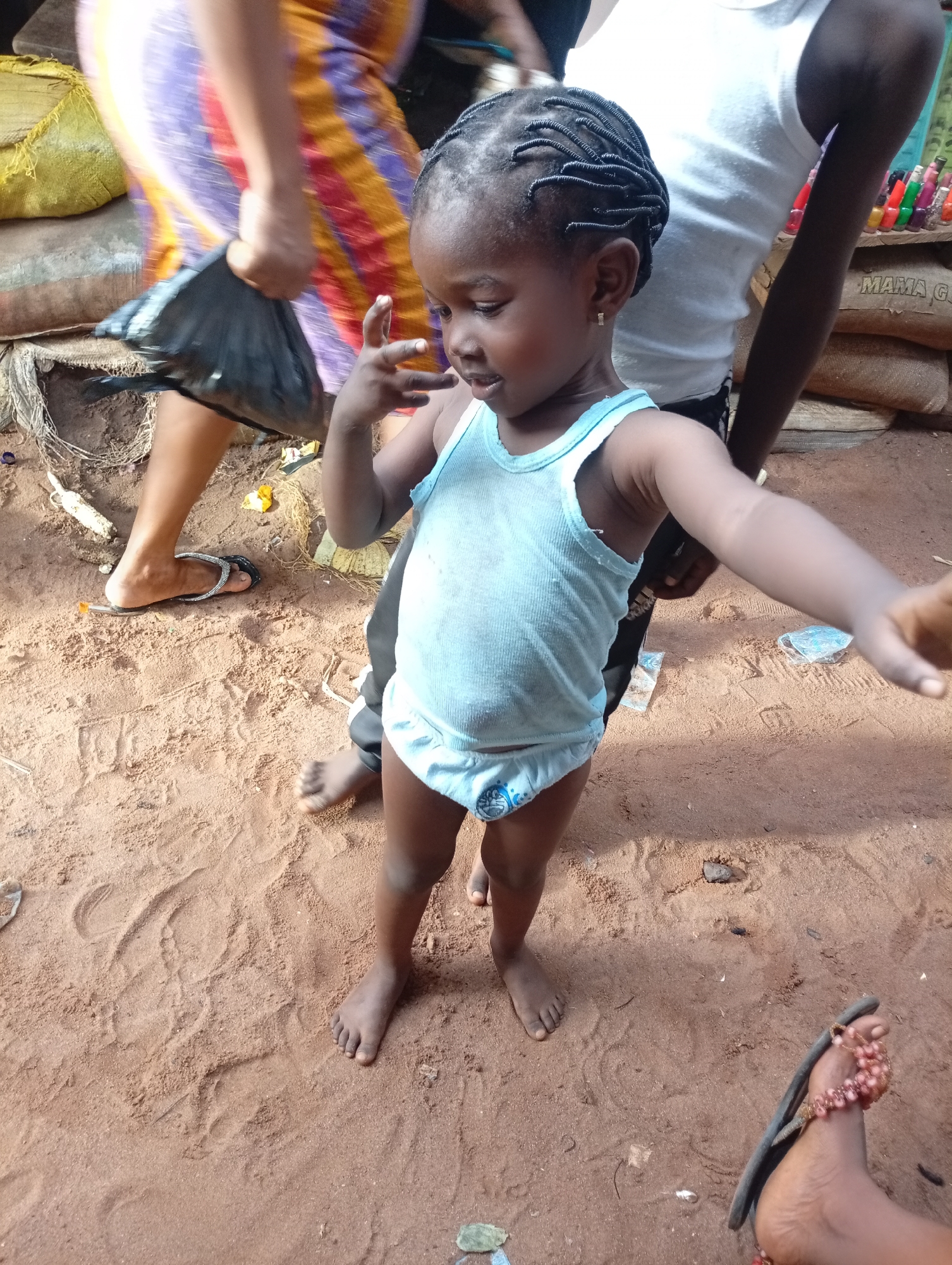 A young Black girl, wearing a light blue tank top and matching underwear with a cartoon character on it, stands on reddish-brown sand. Her hair is styled in numerous small braids. She is looking down and to her right, with her left hand held up and fingers spread, as if reaching for something. Her right arm is extended outwards, towards the viewer.
Behind her, the legs of at least two other people are visible. One person on the left is wearing a colorful, striped wrap skirt and carrying a black plastic bag. Another person behind the girl is wearing a white tank top and dark pants. A shelf with colorful bottles, possibly cosmetic products, is visible behind them, along with a sack labeled "MAMA G". The ground is sandy with some scattered debris, including bits of string and plastic. The scene appears to be outdoors, possibly a market or a street. The lighting suggests it is daytime. The girl's expression is not clearly discernible due to her downward gaze, but her posture and extended arm might indicate curiosity or an attempt to interact.