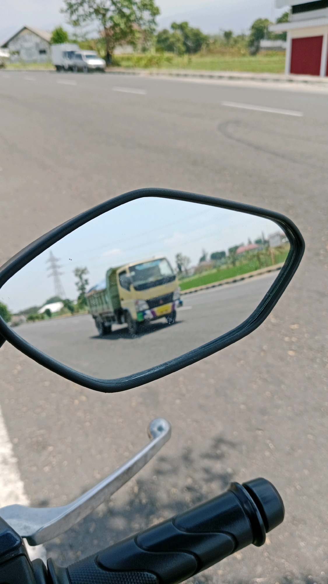 A motorcycle's side-view mirror reflects a yellow and green truck driving on a paved road. The truck is in motion, and its license plate is visible. In the background, a white building and a power pylon are discernible. The scene appears to be a rural or suburban area in Sukowinangun, Indonesia, given the presence of the truck and the road infrastructure. The motorcycle's handlebars are in the foreground, indicating the perspective of the photographer.