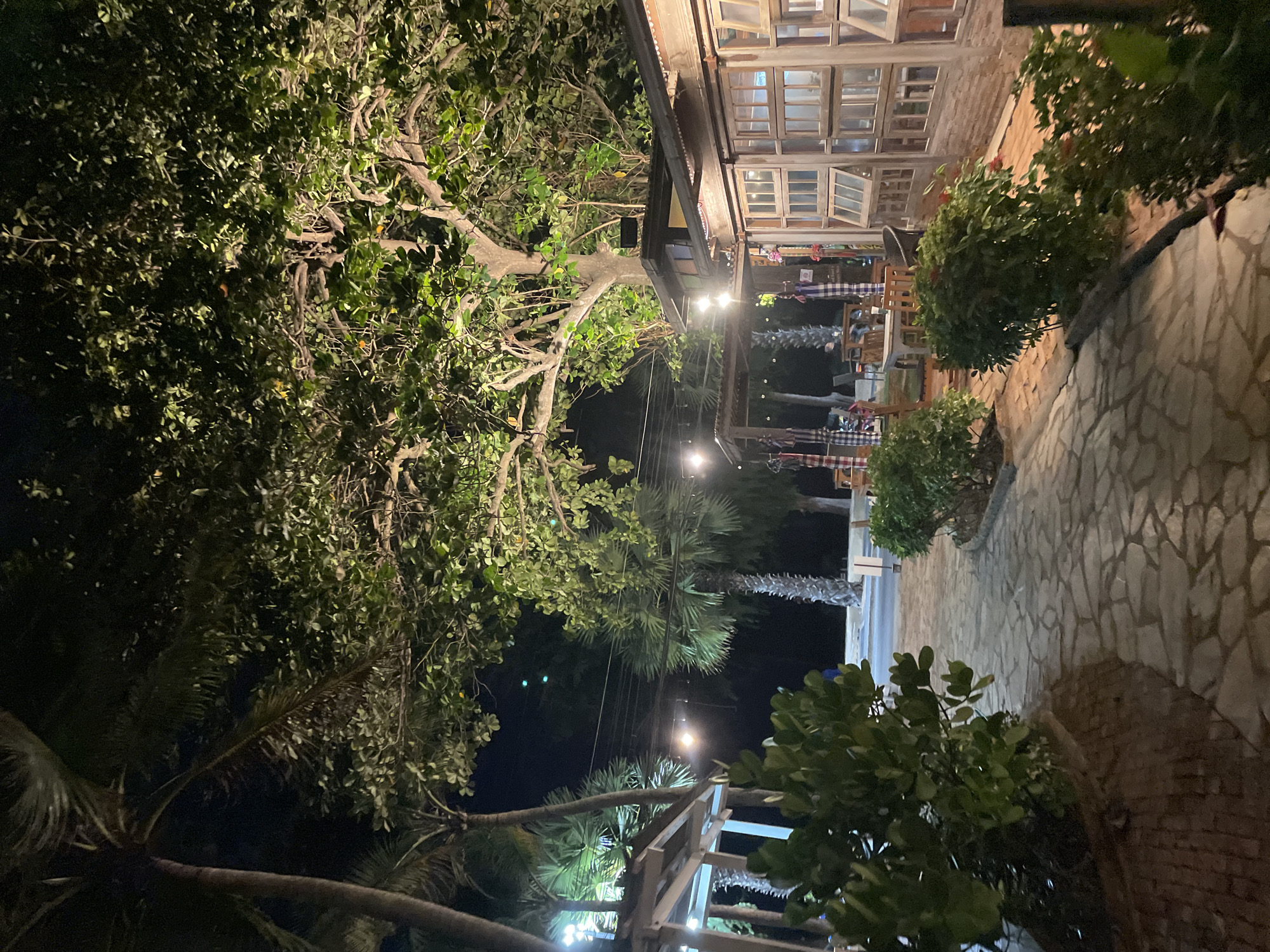 The image depicts an outdoor seating area at night, likely a restaurant or bar, in Pattaya City, Thailand. The scene is illuminated by string lights and ambient lighting from nearby structures. A cobblestone pathway leads through the foreground, flanked by manicured bushes on either side. To the left, a covered structure with white pillars and a sloped roof is visible, suggesting an open-air dining or seating area. Palm trees and other lush foliage are abundant, contributing to a tropical ambiance. To the right, a building with a rustic facade features multiple large windows, some of which appear to be open or have shutters. In the background, tables and chairs are set up under a canopy, with a few patrons visible. The overall atmosphere is relaxed and inviting, characteristic of a nighttime establishment in a beachside city.