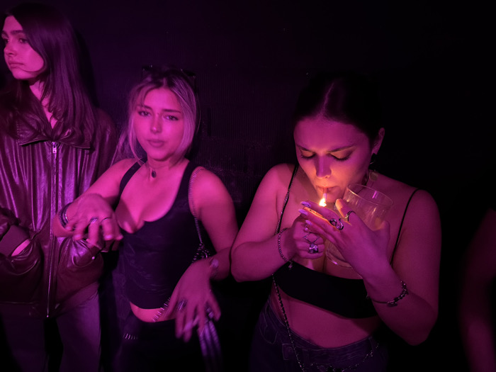 The image displays three young adult females within a dimly lit indoor environment, consistent with a nightclub or bar in Barcelona, Spain. Predominantly illuminated by purple-pink ambient light, the background is dark and indistinct. On the far left, a female with long dark hair, wearing a dark, possibly leather, jacket, is visible in profile, looking towards the left. The central figure is a blonde-haired female with sunglasses perched on her head. She wears a dark tank top, multiple rings on her fingers, and holds an unlit cigarette in her right hand while looking directly forward. The rightmost figure, a female with dark hair pulled back, is actively igniting a cigarette held in her mouth using a lighter in her right hand. Her face is illuminated by the bright orange-yellow flame of the lighter, contrasting with the ambient lighting. She holds a clear glass in her left hand and wears a dark strap top, numerous rings, and a bracelet on her left wrist.