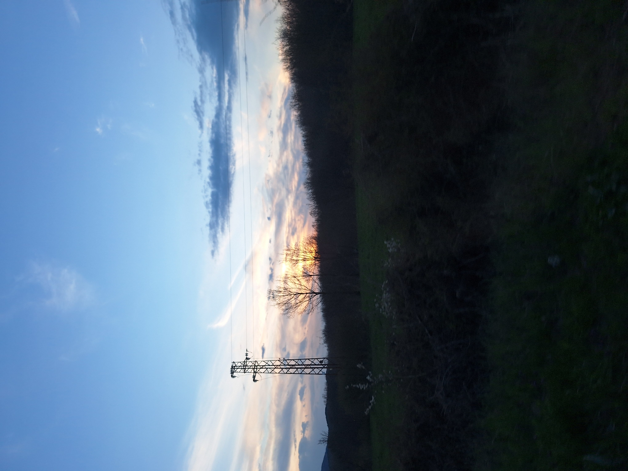 The image depicts an outdoor scene at dusk, likely in a rural or semi-rural area near Batoshevo, Bulgaria. A metal electricity pylon is silhouetted against a sky filled with light blue, white, and dusky pink clouds. Power lines stretch from the pylon into the distance. To the right of the pylon, there is a line of vegetation with trees and green grass. The vegetation is in varying degrees of light and shadow, possibly due to the setting sun. A bare tree stands silhouetted against the sky. The bottom of the image reveals the top of a hill. The sky is the most prominent feature, making up roughly half of the image.