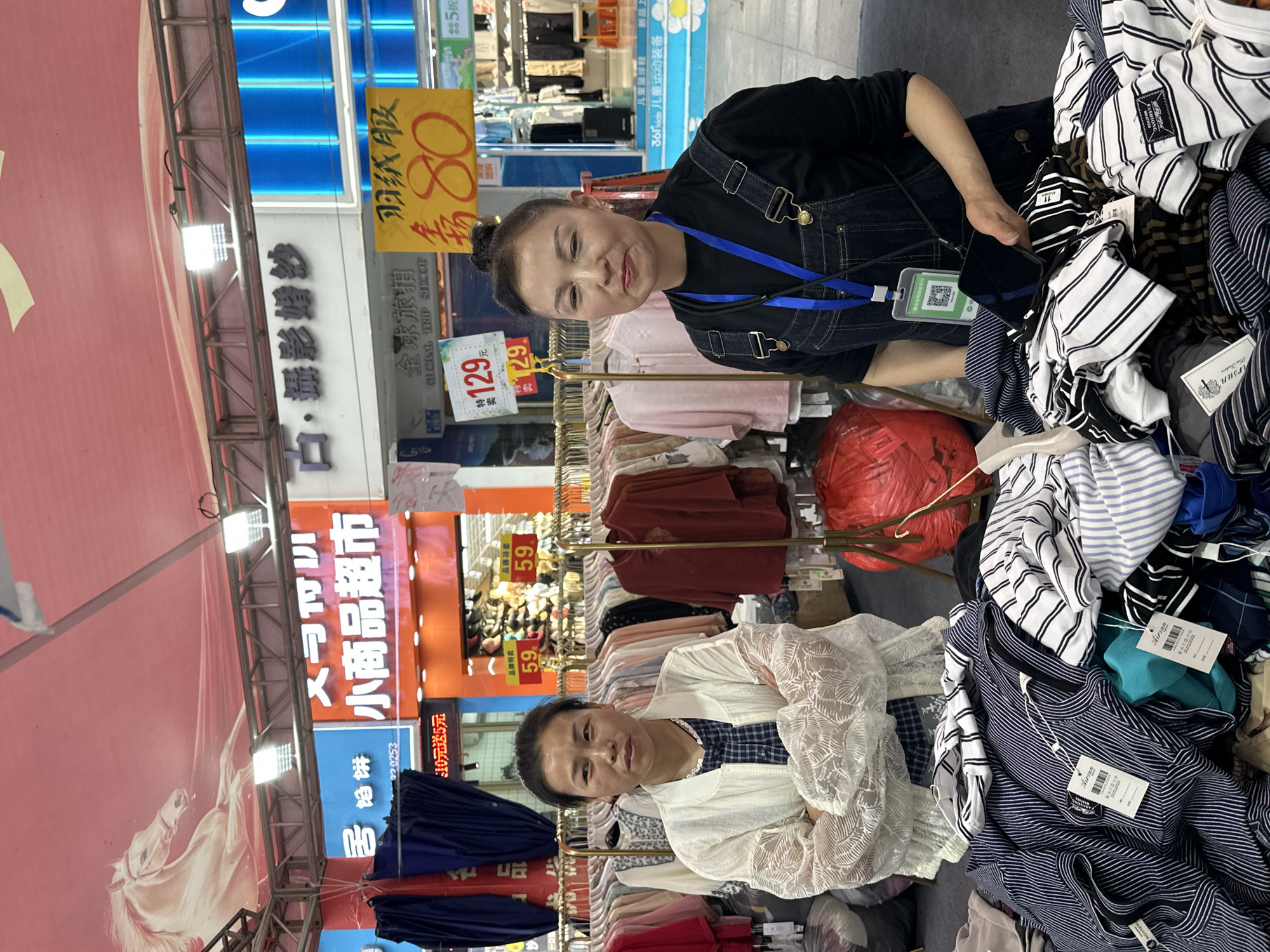 Two women are in a retail setting in Jiangjin, China. One woman, wearing a black shirt and overalls, holds a smartphone near a pile of striped shirts with tags from the "Airsun" brand. The other woman stands by a rack of clothing, wearing a white, lacey shirt and folding her arms. Overhead, there are signs advertising items and prices, including a sign for "全球旅拍" ("Global Trip Shoot") and another for "小商品超市" ("Small Commodities Supermarket"). There's also a promotional banner for "儿童篮球鞋" ("Children's Basketball Shoes"). The background shows other items for sale, including clothing and other products on shelves.