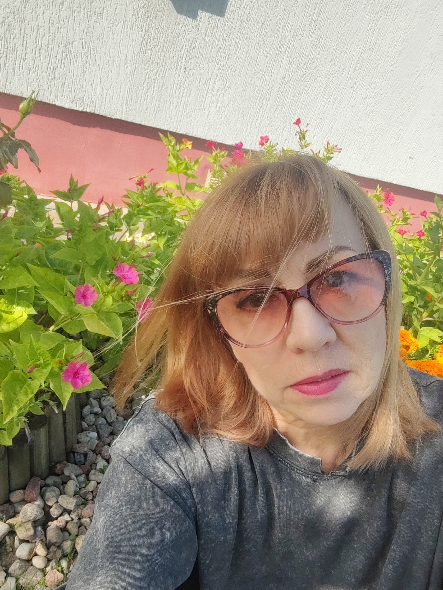 A woman with blonde hair and sunglasses takes a selfie in front of a wall and flowers. The wall is white with a pink band below it. In front of the wall are pink and orange flowers with green leaves. The woman is wearing a dark grey t-shirt. The ground is covered in stones. The scene appears to be outdoors, possibly in a garden or yard.