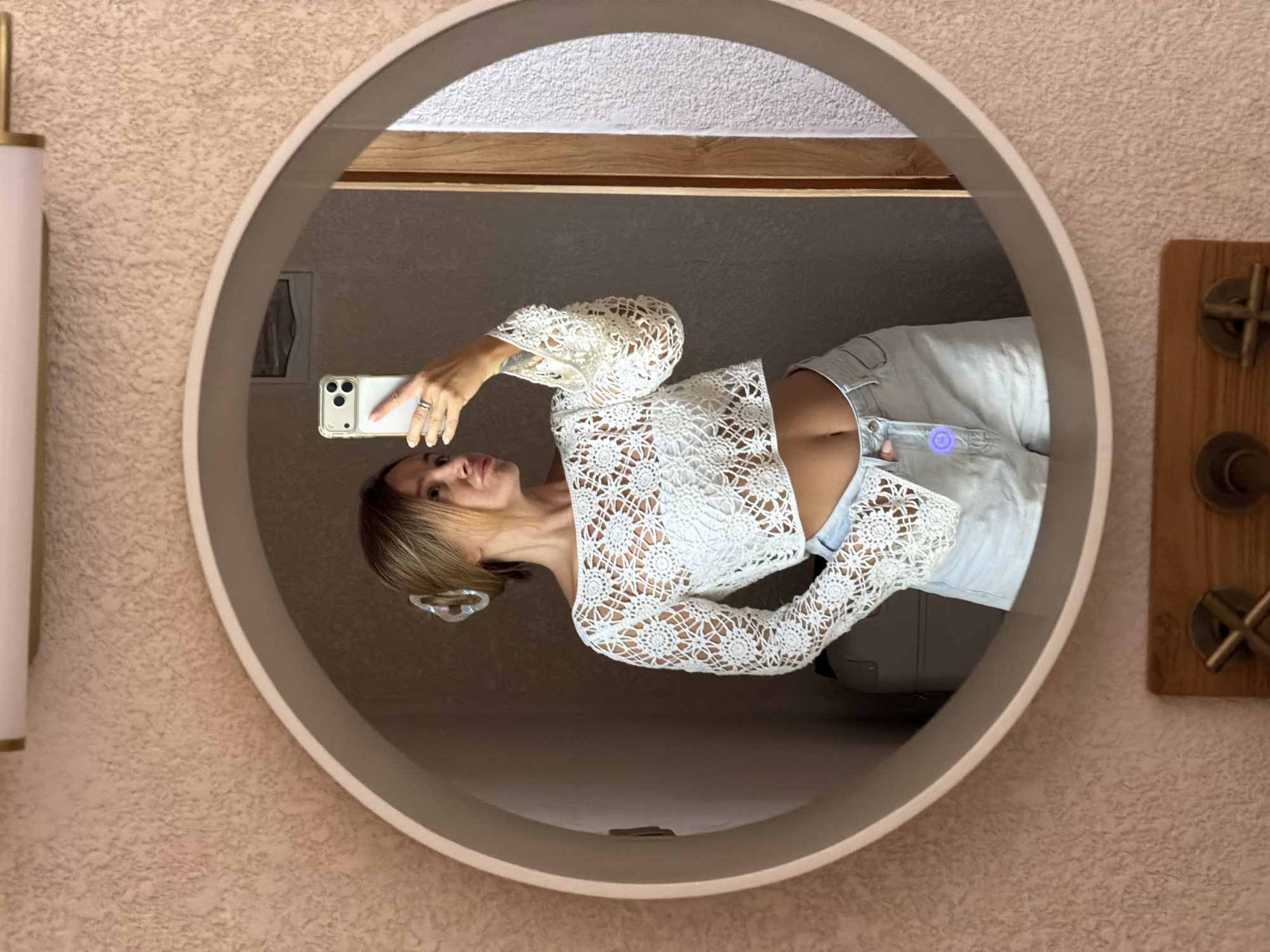 A woman is taking a mirror selfie in a bathroom. She is wearing a white crochet crop top and light blue denim shorts. Her hair is pulled back in a bun and she has a hair clip in it. The bathroom has pink textured walls and a round mirror. There is a light fixture above the mirror.