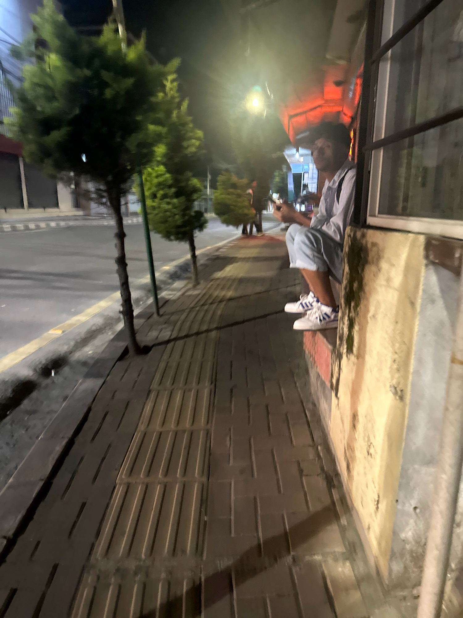 A person sits on a ledge outside a building at night in Lalitpur, Nepal. The individual is wearing a light-colored shirt, light blue shorts, and white sneakers with blue stripes. They are holding a mobile phone. The ledge appears to be part of an establishment with a window on the right and an awning with red lighting above. In the background, a street with trees and streetlights can be seen. The ground in front of the ledge is paved with tactile paving bricks, commonly found on sidewalks for visually impaired pedestrians. The street is wet, suggesting recent rain.