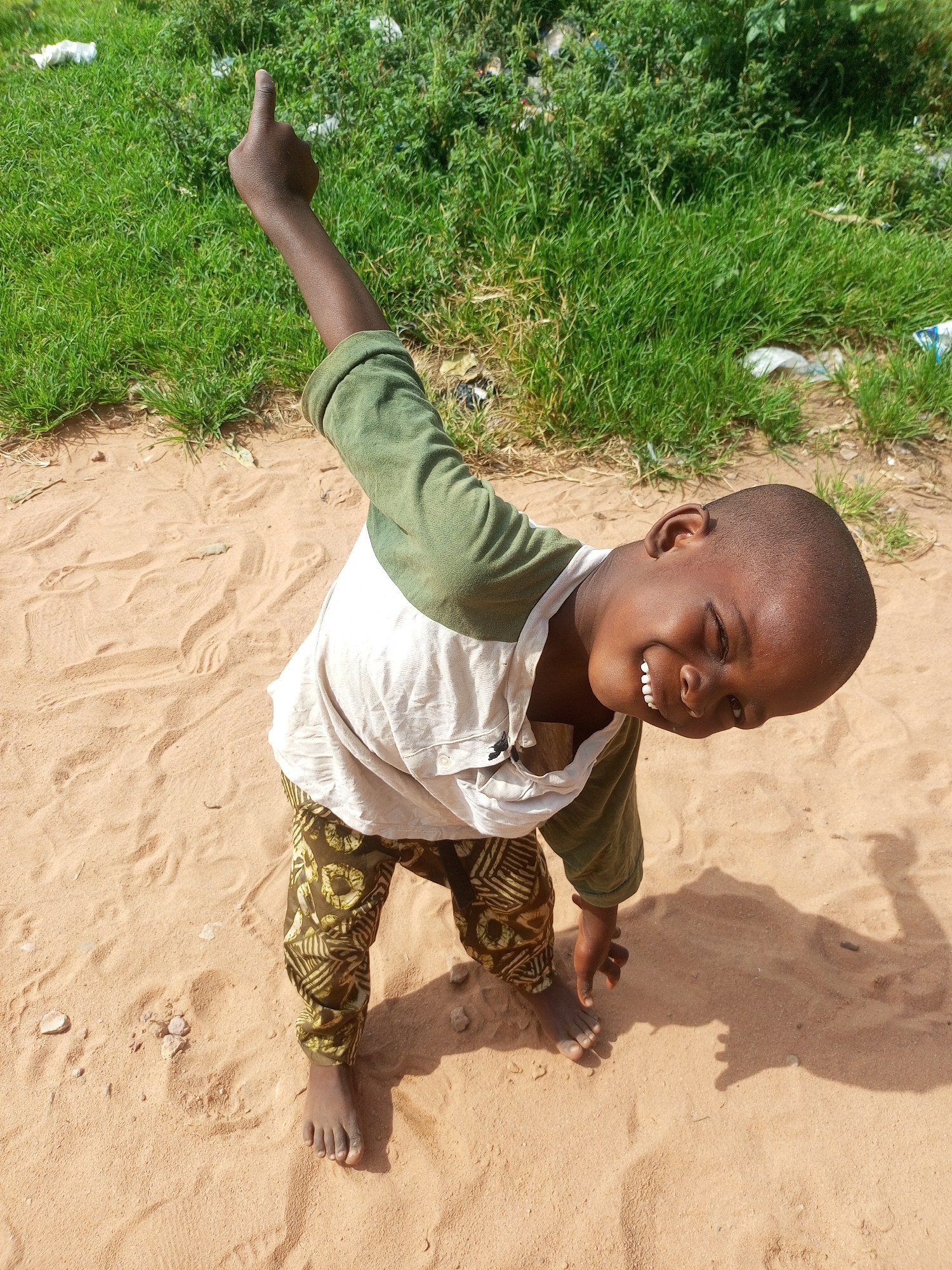 A young boy with dark skin and short hair is pictured in a sandy area with green grass and debris in the background. He is wearing a green long-sleeved shirt over a white t-shirt and patterned pants. His head is tilted back, and he is smiling with his eyes closed. His right arm is raised, pointing upwards with his index finger. The environment suggests an outdoor setting in a city like Tempure, Nigeria. The ground is sandy with some footprints and small stones. The background features a patch of green grass with some scattered white plastic bags. The lighting indicates it is daytime.