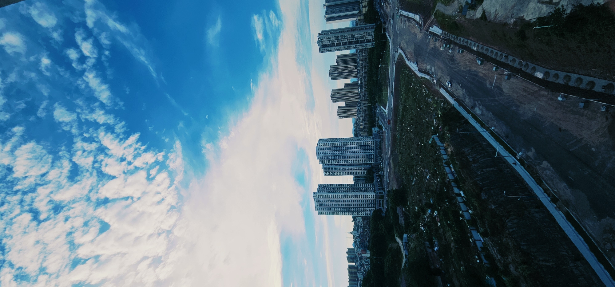 An aerial, vertical shot depicts a modern cityscape under a bright blue sky dotted with white clouds. In the foreground, a developing area features a paved road curving through undeveloped land with patches of green vegetation and some debris. Several lamp posts line the road. Beyond this, a densely populated residential area is visible, characterized by numerous tall, modern apartment buildings with a uniform architectural style. The buildings are white or light-colored and appear to be arranged in clusters. Further in the distance, a more traditional urban landscape with lower-rise buildings can be seen. The image appears to be taken from a high vantage point, possibly a drone or tall building, looking down and across the city. This scene is located in Jiulongpo District, China.