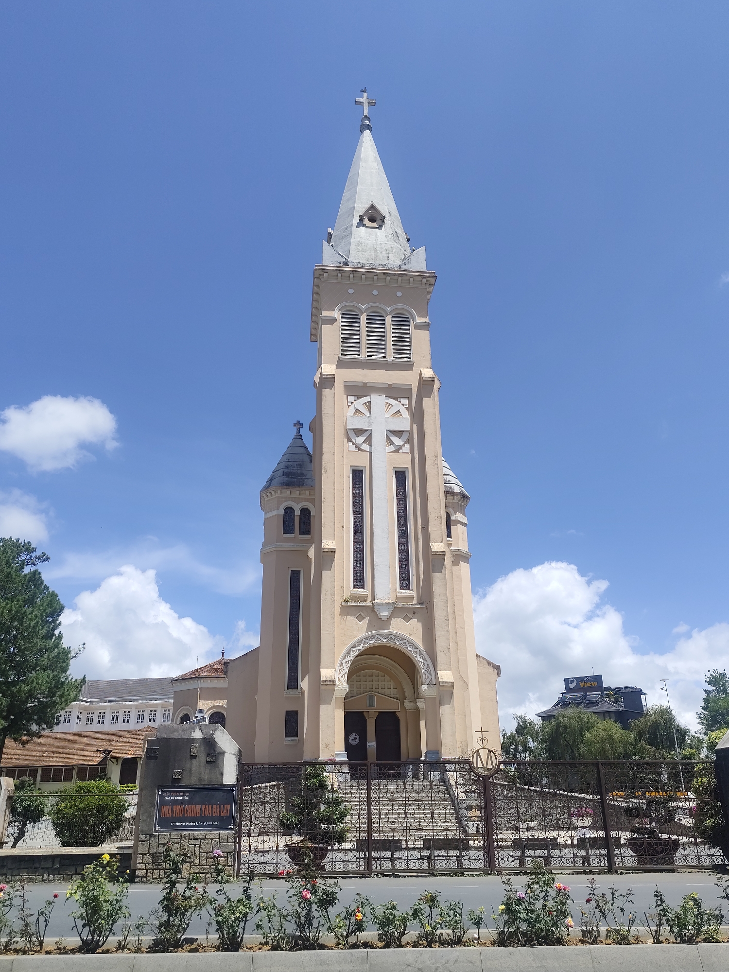 This is a front-facing, eye-level, full shot of the Da Lat Cathedral in Da Lat, Vietnam. The cathedral, a Roman Catholic church, is made of beige stone and features a tall bell tower topped with a cross. The tower has arched windows and a large white cross on its facade. The cathedral is set behind a decorative metal fence and a flowerbed. In the background, a blue sky with scattered white clouds is visible, along with some trees and other buildings. The overall impression is one of a well-maintained, historic religious building in an urban setting.
