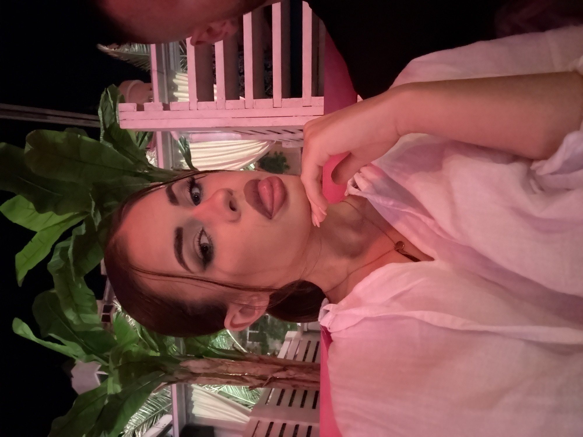 The image presents a playful, close-up portrait of a young woman in Plovdiv, Bulgaria, captured during the evening or night. She is positioned with her head tilted to the side, looking directly at the camera. Her expression is distinctive, with her tongue slightly out and shaped into a heart. She has dark hair, styled in an updo, and prominent eye makeup accentuating her light-colored eyes. Her right hand is visible, with manicured nails, gently touching her chin. She wears a loose-fitting, light-colored (possibly white or pale pink) top and a delicate gold-toned necklace around her neck.
The background is dimly lit with a strong pinkish-red artificial light, creating a warm, moody atmosphere. Large, dark green, palm-like leaves are visible, suggesting tropical or decorative foliage. A white lattice-like structure, possibly a trellis or decorative partition, and other subtle architectural elements are also present. These background details hint at an outdoor or semi-outdoor venue, such as a restaurant or lounge. A small section of another person's head is visible at the very top edge of the frame. There is no visible text in the image.
