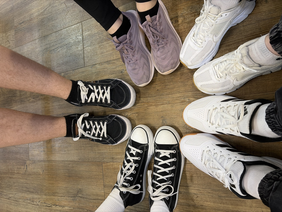The image displays the feet and lower legs of six individuals arranged in a rough circular formation on a light brown wooden floor, located in the city of Chaikovsky, Russia.
From the top and moving clockwise:
1. One individual presents with bare, hairy lower legs, wearing black canvas low-top sneakers with white laces and black ankle socks.
2. One individual wears black trousers or leggings, light purple mesh athletic shoes with matching laces, and black ankle socks.
3. One individual wears white mesh athletic shoes with white laces and white ankle socks.
4. One individual wears dark cuffed trousers, white athletic shoes with black stripes on the sides, white laces, and white ankle socks.
5. One individual wears black canvas high-top sneakers with white laces, white soles, and white ankle socks.
6. One individual wears black canvas high-top sneakers with white laces, white soles, and white ankle socks, positioned slightly above the fifth individual's feet.
The group's feet are pointed towards the approximate center of the circle, where the individual with bare, hairy legs is located at the apex of the circle. The wooden floor exhibits a plank or laminate pattern.