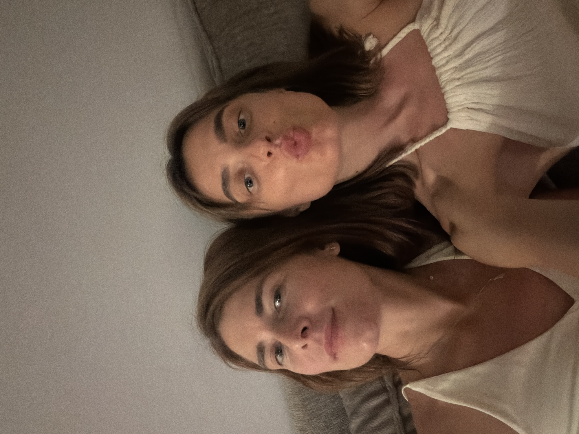 Two women are captured in a selfie. The woman on the left has light brown hair and is looking directly at the camera with a neutral expression. She is wearing a white top with thin straps. The woman on the right has light brown hair and is puckering her lips as if to blow a kiss. She is also wearing a white top with similar thin straps. They appear to be indoors, possibly on a couch or bed, with a textured grey background that could be a wall or furniture. The lighting is dim, suggesting it is evening or nighttime. There are no visible landmarks or clues to indicate the specific location within Tel-Aviv, Israel.
