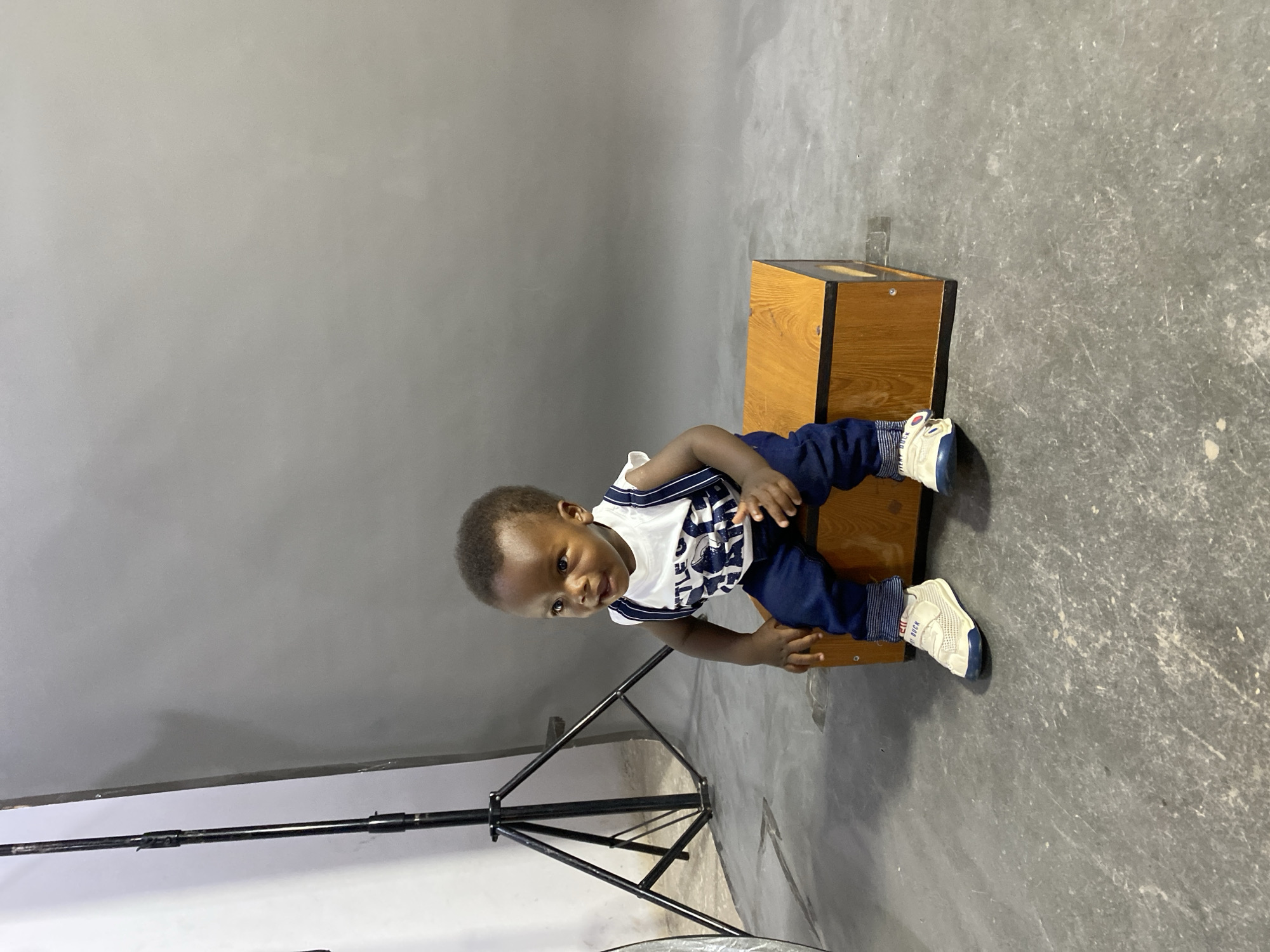 The image displays a young child, likely a baby or toddler, posing in what appears to be a photography studio in Gura Top, Nigeria. The child is positioned on a light-brown wooden box with dark edges, sitting or kneeling, and looking directly at the camera with a curious, gentle smile.
The child is dressed in a white sleeveless top with blue stripes and some partially visible text, dark blue pants, and white and blue sneakers. On the child's right shoe, the text "HI DUCK" is clearly visible.
The background is a simple gray wall or backdrop, transitioning to a grayer, textured concrete floor below. To the left of the child, a black photography light stand or tripod is visible, along with part of a circular photography reflector on the floor. The studio setting suggests a planned photoshoot. The child appears happy and engaged.
