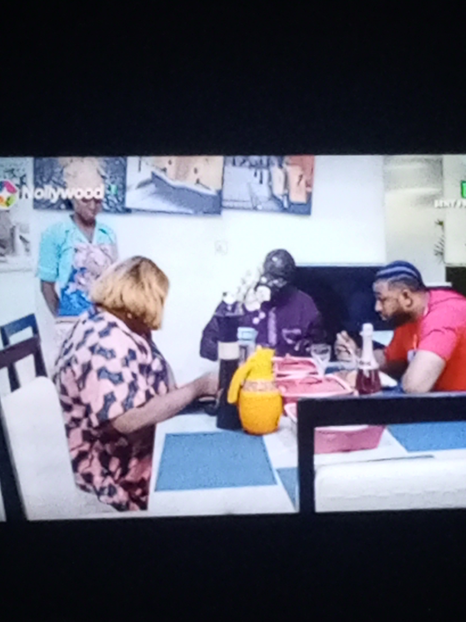 Here is a forensic description of the image:
The image depicts an indoor scene involving four individuals seated around a table. The table setting includes plates, glasses, and a yellow pitcher. To the left, a person with blonde hair, wearing a pink and black patterned shirt, sits with their back to the camera. Behind them, a woman in a light blue shirt stands with her hands behind her back. Across the table, an individual in a purple shirt is visible, engaged in what appears to be a phone call. To their right, another person wearing a pink shirt with blue braids sits at the table.
The location appears to be a dining area, with artwork hanging on the wall in the background. A television channel logo, "Hollywood," is visible in the top left corner of the image. The city of Unknown, Unknown is not identifiable from the given visual information.