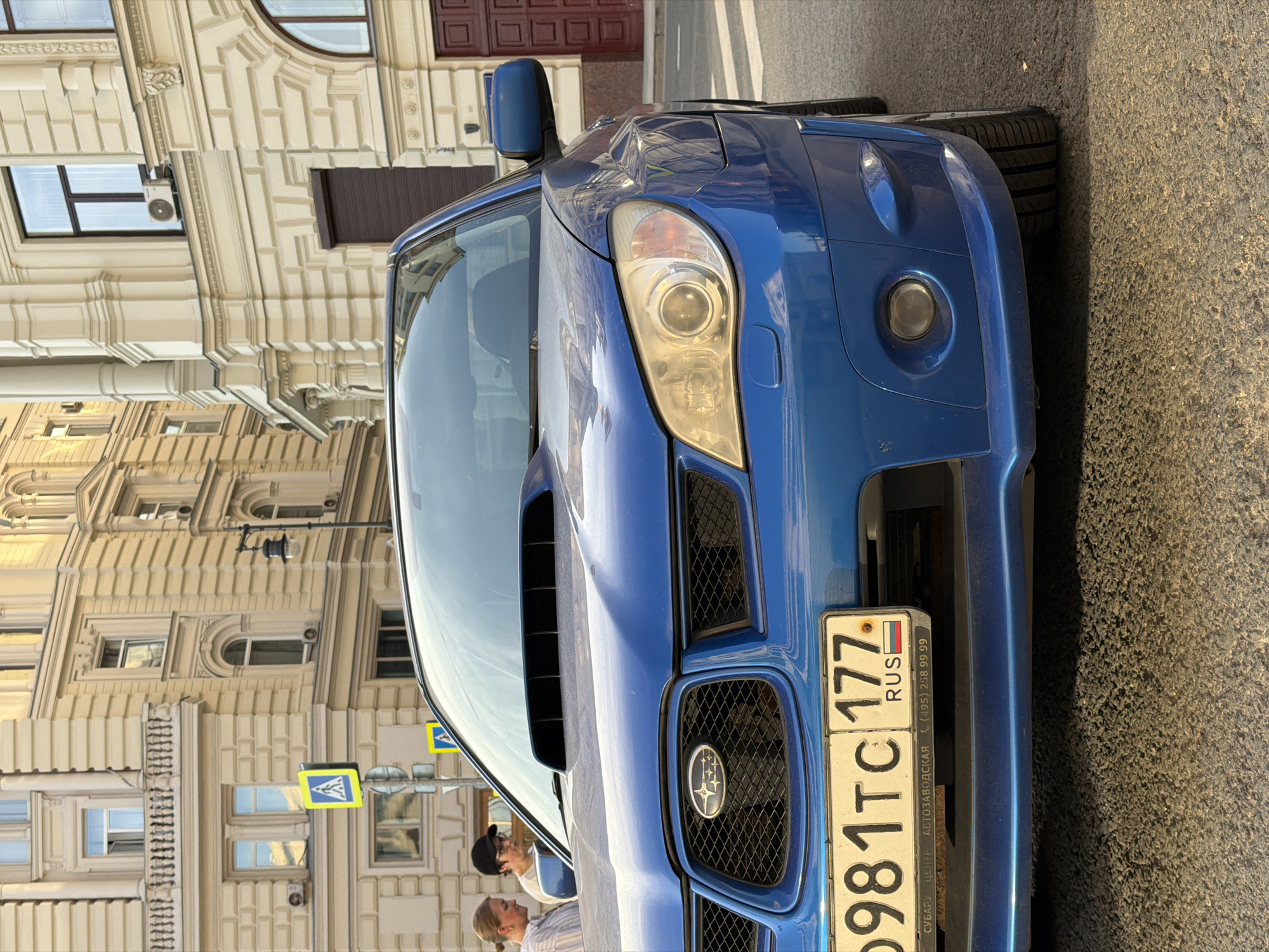 The image shows the front of a blue Subaru Impreza WRX parked on a street. The car has a Russian license plate that reads "O981TC 177 RUS". The background features a building with detailed architectural elements and a pedestrian crossing sign. Two people are standing near the car. The location is Moscow, Russia, based on the license plate and architectural style of the building. The image is rotated counter-clockwise.