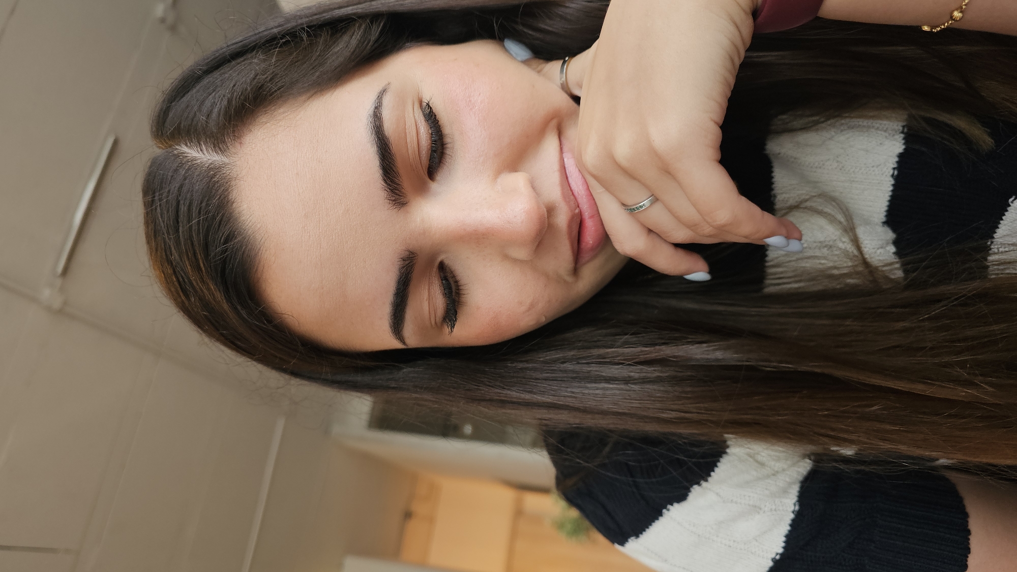 A young woman with long brown hair, wearing a black and white striped sweater, is resting her head on her hand. She has fair skin, closed eyes, and a slight smile. She is wearing a ring on her finger. The background is blurred, suggesting an interior setting, possibly a home. There is no indication of Puebla, Mexico.