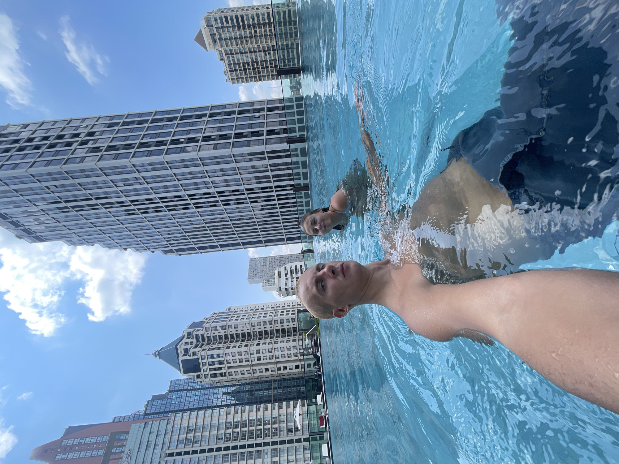 Two individuals are captured in an outdoor swimming pool, likely situated atop a tall building in Bangkok, Thailand, evidenced by the numerous high-rise buildings visible in the background. The buildings exhibit a variety of architectural styles and window patterns. The foreground shows the first individual, who appears to be fair-skinned with short blonde hair, with the water level at their chest. The second individual is slightly behind. The water is light blue and there are ripples on the surface. The weather is clear and sunny with some clouds.