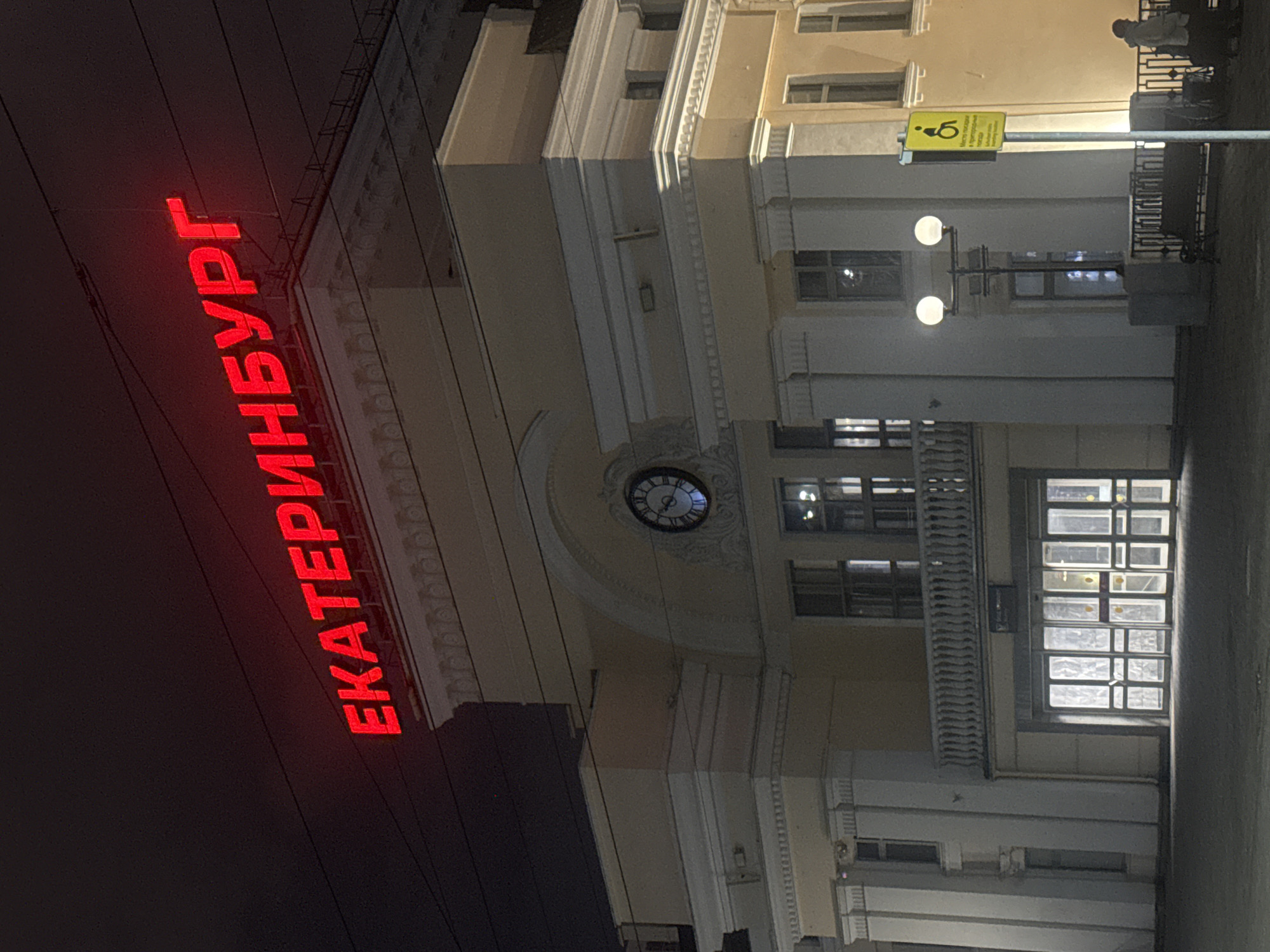 The image depicts the Yekaterinburg railway station at night. The façade of the building is visible, featuring a clock and rows of windows. A sign with the name of the city, "ЕКАТЕРИНБУРГ," is illuminated in red. A street lamp with a disability access sign is also visible.