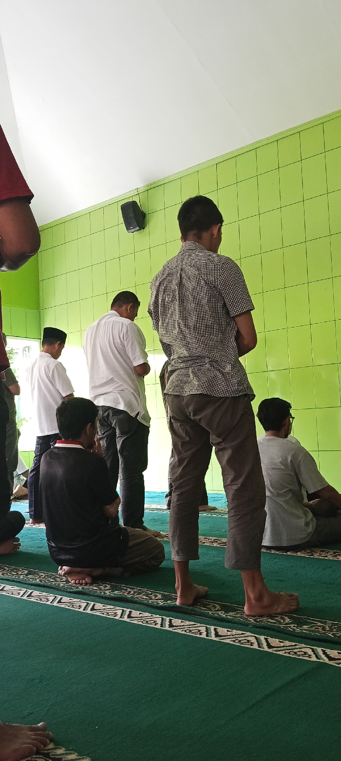 The image shows a group of five men praying inside a mosque in Soreang, Indonesia, located at the coordinates -7.022, 107.524.  The men are dressed in traditional Muslim clothing. They are facing a wall with green tiles.  There is a dark speaker attached to the wall.  A patterned green rug covers the floor, with a white border. The man in the center is the most visible, with his back to the camera. He is wearing a plaid shirt and brown pants. The other men are partially visible, with their heads bowed in prayer.  The room is lit by natural light, and the atmosphere is peaceful and solemn. The scene suggests a routine Islamic ritual taking place.