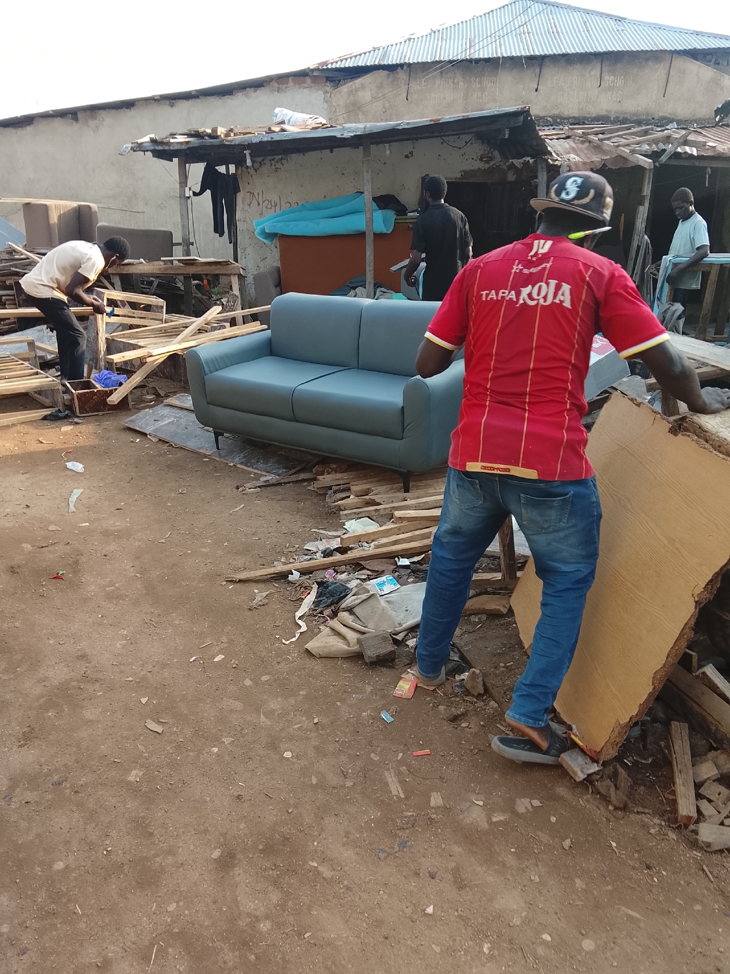 In the city of Abuja, Nigeria, a scene of furniture assembly or repair unfolds in an outdoor workshop setting. The environment is characterized by its rustic, unpaved ground, littered with wood scraps, debris, and what appears to be discarded materials. A corrugated metal roof, supported by makeshift wooden structures, provides partial overhead cover.
In the foreground, a person wearing a red shirt with yellow and white stripes and blue jeans is actively engaged in moving a large, flat piece of wood, possibly a slab or a tabletop. They are wearing dark sandals. Behind them, a light blue, modern-looking sofa is positioned on wooden planks and a dark tarp.
To the left of the sofa, another individual in a light-colored shirt and dark pants is seen working on wooden furniture frames with tools, suggesting construction or refurbishment. Further back, two other men are visible. One is standing near a blue tarp, and another is looking on from behind a wooden structure. The overall impression is one of a busy, informal workshop where furniture is being crafted or repurposed amidst a somewhat disorganized but functional outdoor space.
