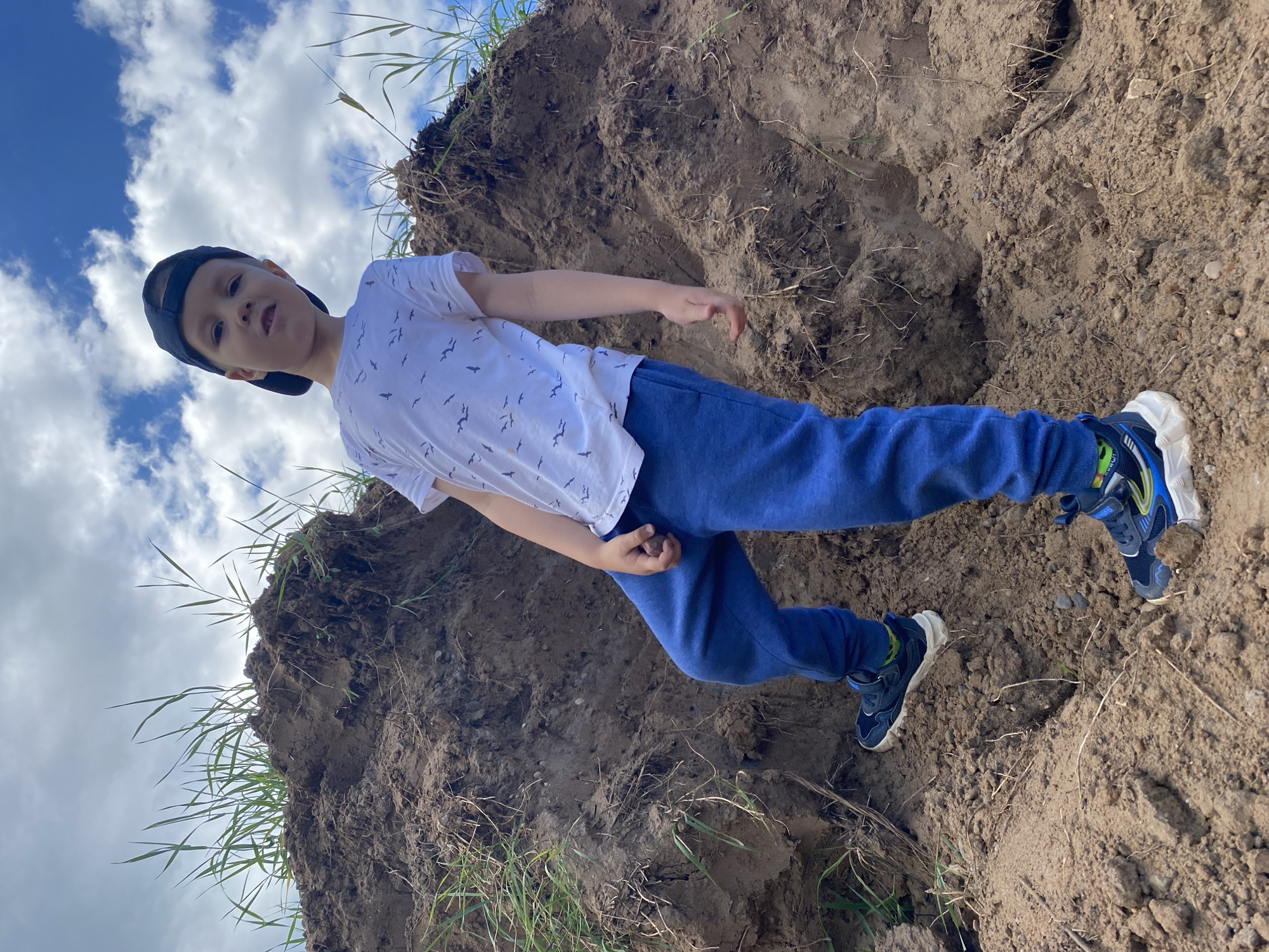 The image, taken in Chaikovsky, Russia, depicts a young Caucasian male child standing on a large mound of loose, brown earth. The perspective is low-angle, looking up at the subject and the sky. The child wears a dark blue baseball cap, a white t-shirt patterned with black bird silhouettes, blue sweatpants, and multi-colored athletic shoes with prominent white soles (dark blue, white, light blue, yellow, and green accents). His right foot is positioned higher on the mound, and he makes direct eye contact with the camera, his expression neutral. He holds a small, dark rock or clod of dirt in his right hand. Sparse green grass blades and dried roots protrude from the dirt mound. The background shows a partially cloudy sky with visible patches of blue.