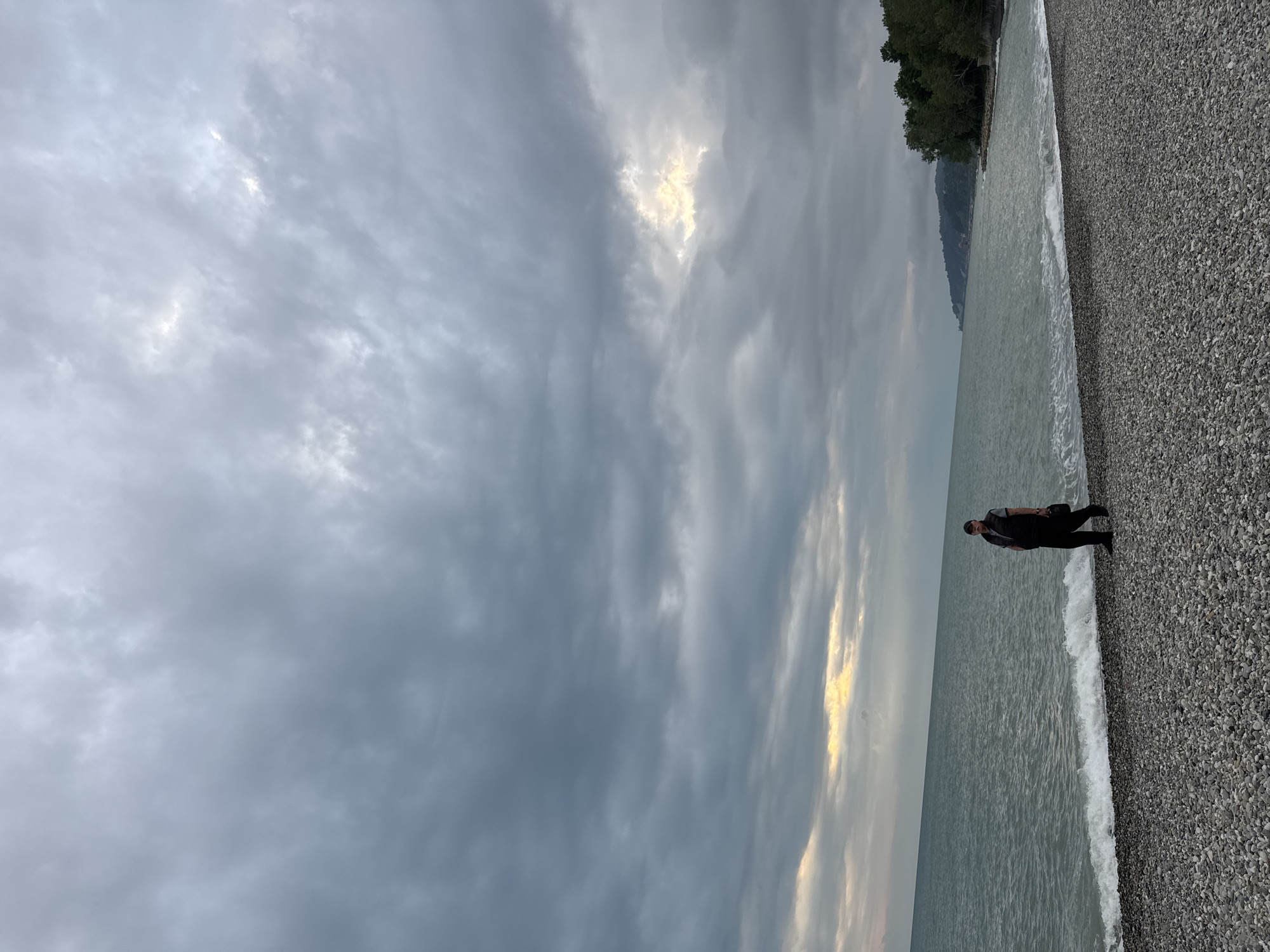 A person is standing on a pebble beach next to the water. The sky is overcast with a break in the clouds revealing sunlight. Trees are visible in the distance along the coast. Based on the geography and details such as the pebble beach, the location is likely in Sochi, Russia.
