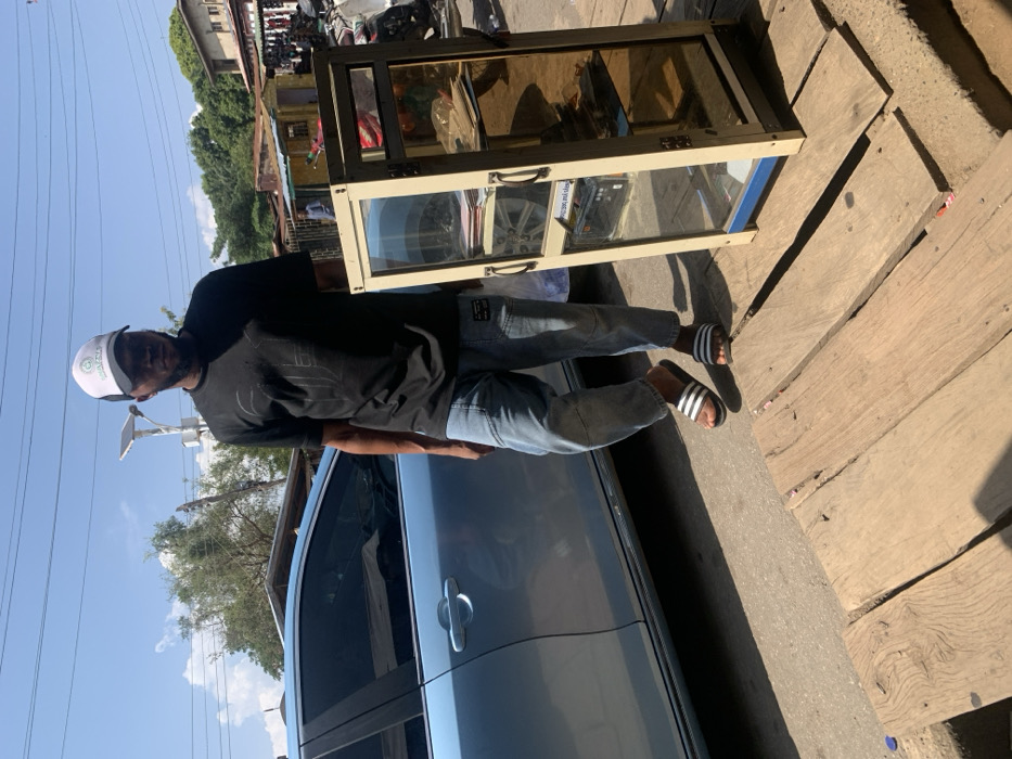 This image depicts an outdoor street scene in Okene, Nigeria, characterized by bright daylight and commercial activity. The primary subject is a male individual, appearing to be of African descent, standing in the midground, oriented slightly to the viewer's right. He wears a white baseball cap with a green circular logo, a black short-sleeved t-shirt with a dark graphic, light blue denim jeans, and black-and-white striped slide sandals. He has a beard.
To his immediate right stands a light-colored display cabinet, approximately waist-height, constructed with a metal frame (likely aluminum) and glass panels, including some with a light amber tint. The cabinet contains papers and various items on multiple shelves. A black rectangular object, possibly an electronic device or battery, is visible on a lower shelf behind a clear glass panel. The cabinet rests on a platform composed of several rough, unpainted wooden planks.
Behind the man and to his left, a light blue sedan car, possibly a Toyota, is parked or stationary. The car's side profile, including tinted rear windows and a front door handle, is visible. The ground beneath the man and extending into the foreground consists of these wooden planks, laid over an uneven surface, likely a curb or sidewalk, transitioning to a paved road or concrete surface beyond.
The background reveals a bustling urban environment under a clear blue sky with minimal cloud cover. A utility pole equipped with a solar panel and light fixture is visible behind the man. Beyond the car and the utility pole, a cluster of residential and commercial buildings, some with corrugated metal roofs and painted in various light colors, are visible amidst dense trees and foliage. Further to the right in the background, obscured figures, partial views of motorcycles, and what appear to be additional market stalls or structures are present. Shadows are distinct, indicating strong overhead sunlight.