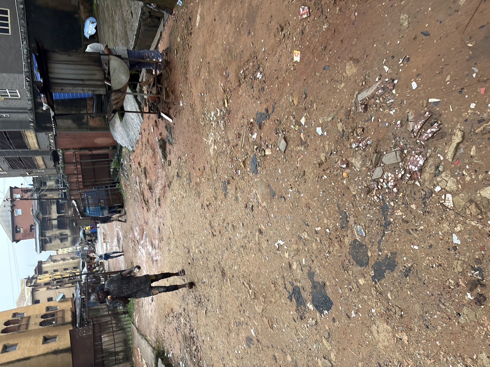 The image depicts a street scene in Onitsha, Nigeria, characterized by a dirt and mud-covered ground, indicative of recent rain or poor drainage. The environment appears somewhat dilapidated, with various buildings lining the street, some appearing aged and weathered. There is a scattering of debris and trash on the ground, contributing to a sense of disarray.
In the foreground, a man wearing a dark, patterned t-shirt and dark pants is walking away from the camera. He appears to be holding something yellow near his mouth. Further down the street, another individual in lighter-colored clothing is visible. To the right, a makeshift food stall is set up, featuring a metal structure with a canopy and a grill. A person is partially visible behind the stall, appearing to be attending to it. Several other individuals are scattered in the background, some near the stall and others walking along the street. A blue truck is parked on the right side of the street. The overall impression is of a busy, working-class urban environment with a challenging infrastructure.