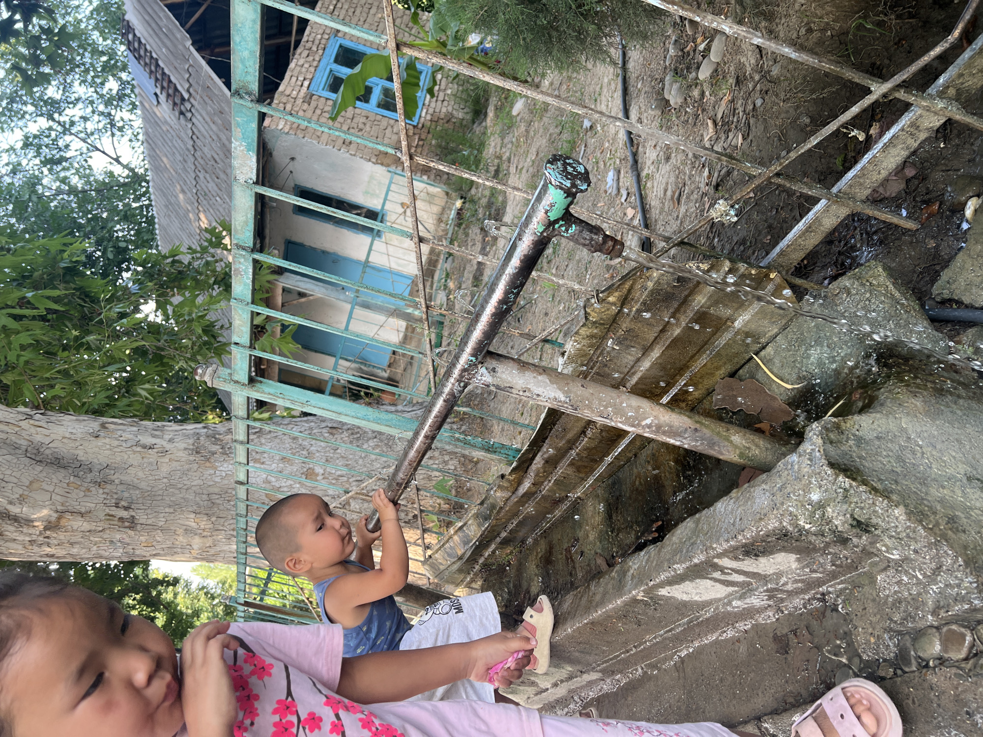 The media file shows two young children near a water source with a metal structure and a building in the background. A baby in a blue tank top is holding onto a metal pipe that is dispensing water. The baby's left hand is on the pipe, and water is flowing from the end of the pipe onto a concrete structure. A young girl is in the foreground, partially out of focus, wearing a pink shirt with floral patterns. Her head is tilted to the side. The background features a weathered building with a blue window and a corrugated roof. Green trees and foliage are visible around the building and to the left of the frame. The scene appears to be outdoors, possibly in a rural or semi-rural setting, given the rustic nature of the building and the presence of an outdoor water source. The ground is a mixture of dirt and concrete. The lighting suggests it is daytime. The overall impression is a candid moment of children interacting with water in a simple environment. No visible text is present in the media file. The location context indicates this scene is in Kara-Dıykan, Kyrgyzstan.