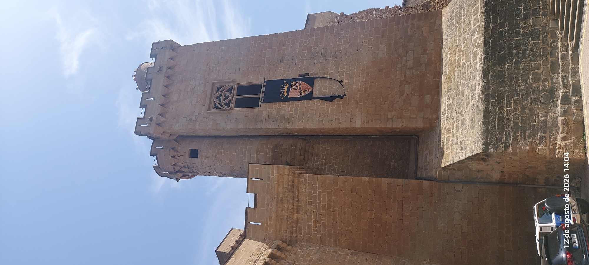 The image depicts a section of the Royal Palace of Olite in Olite / Erriberri, Spain, under a bright, partly cloudy sky. The architecture is characterized by weathered, tan-colored stone blocks forming a large tower with crenellated battlements and a smaller turret to its left. A dark banner with a coat of arms and a crown is affixed to the tower's facade, just below a Gothic-style window. The lower portion of the scene shows a rough-hewn stone ramp leading up to the palace walls. In the bottom left corner, parts of two parked vehicles are visible. The vehicle on the left is black, and the one on the right is a white SUV with a spare tire mounted on the back. The date "12 de agosto de 2026 14:04" is superimposed on the lower left portion of the image, indicating the time and date of capture. Several people are visible on the upper battlements of the tower, appearing as small figures against the sky.