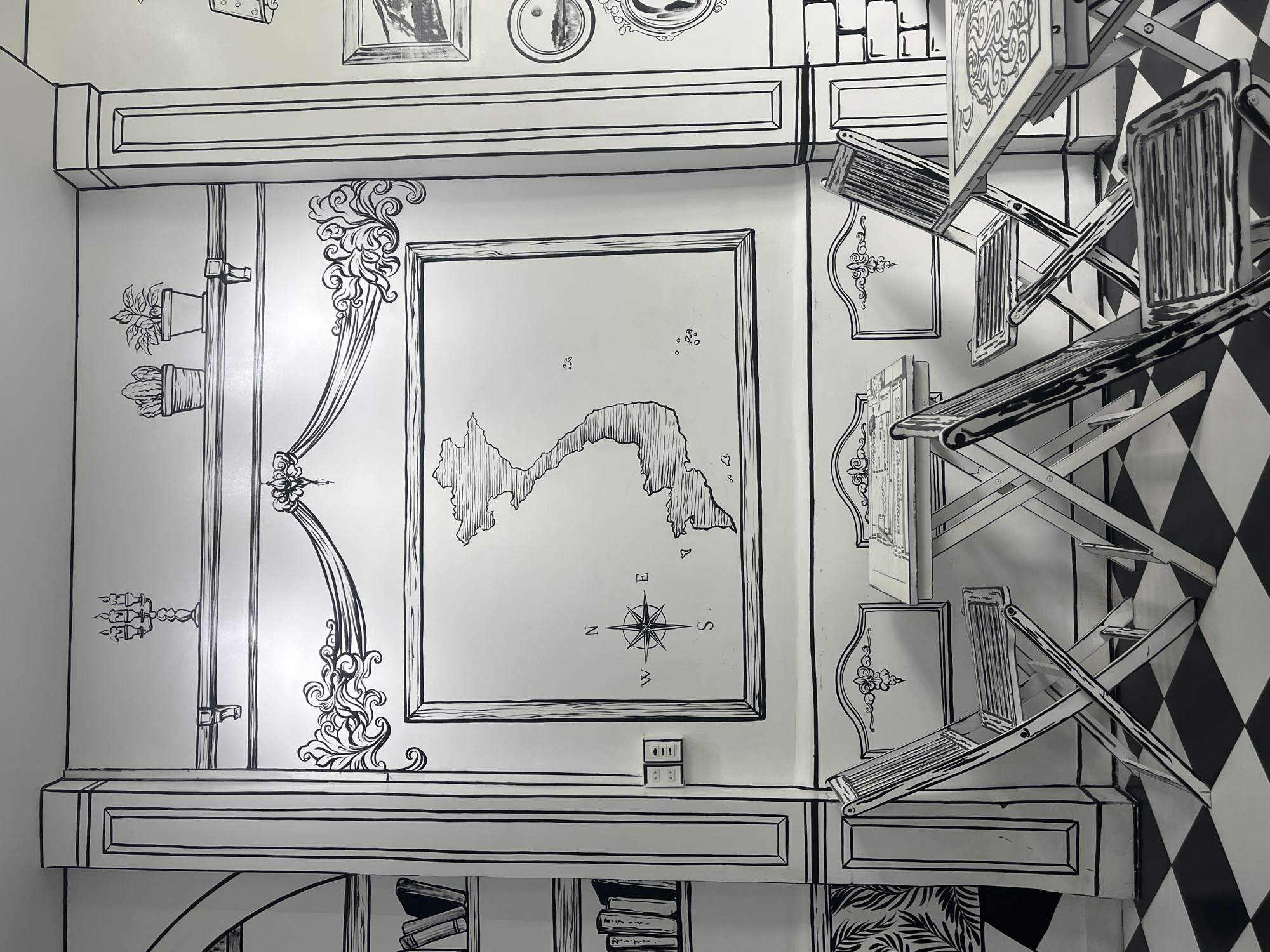 This image depicts the interior of a cafe designed in a two-dimensional, cartoon-like style, resembling a sketch. The walls are white with bold black outlines defining architectural features and decorative elements. On the walls, there are drawings of a candelabra, potted plants, a framed map of Vietnam with a compass rose, and other ornate designs.
In the foreground, there are several folding tables and chairs, also rendered in the same black-and-white sketch style. These furniture pieces have thick black outlines and are arranged on a black and white checkered floor, which further enhances the 2D illusion.
The overall aesthetic suggests a whimsical and artistic environment. While the image itself doesn't explicitly state its location, the presence of a map of Vietnam and its general style are consistent with cafes found in major cities of Vietnam, such as Ho Chi Minh City. There are no people visible in the image, and the cafe appears to be unoccupied at the time the photograph was taken. The lighting is bright and even, highlighting the stark black and white design.