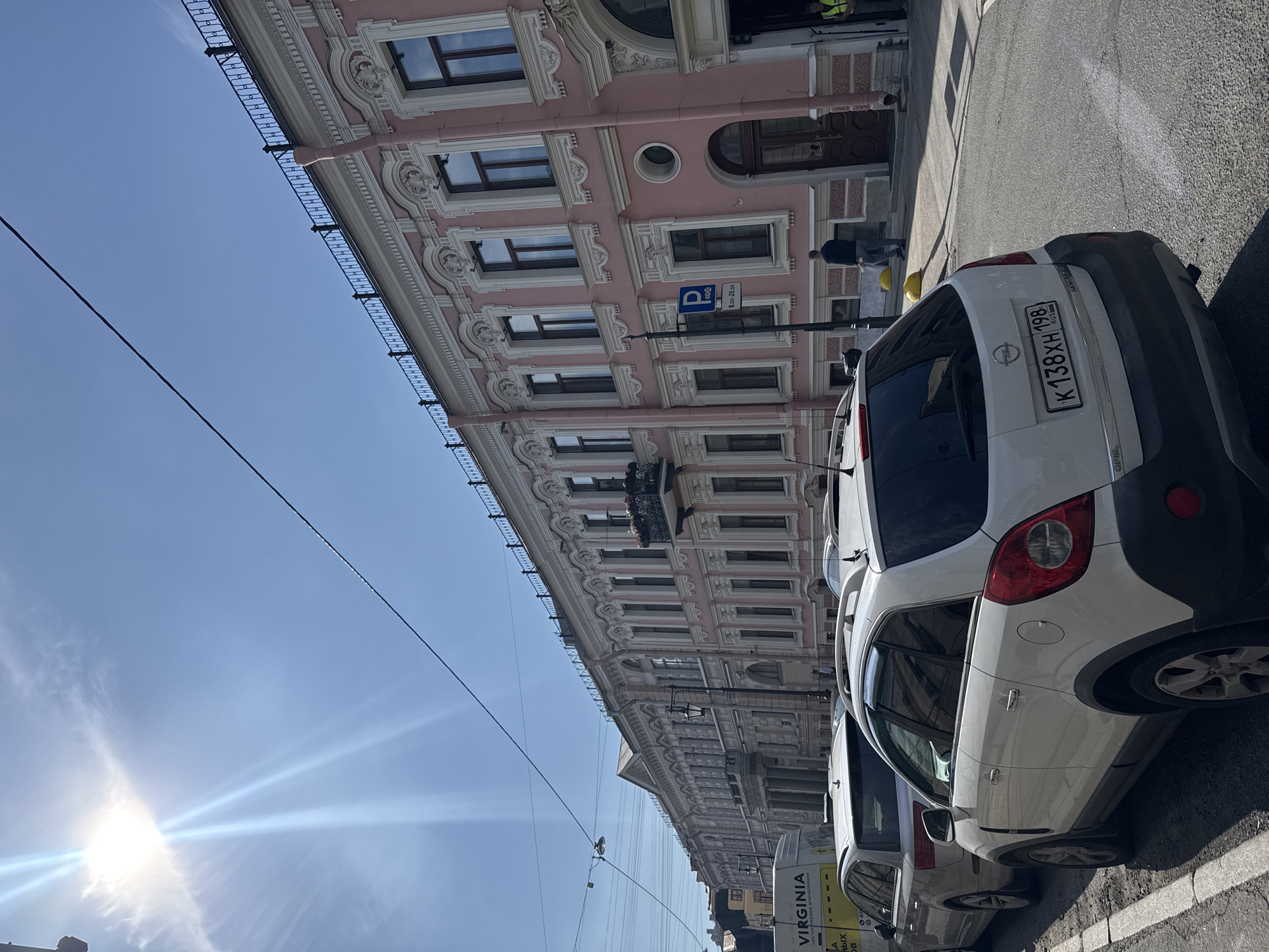 The image depicts a street scene with a pink building in the background and cars parked along the side of the road. The building features ornate architectural details, including columns, decorative moldings, and multiple windows. The sky is a clear blue, suggesting fair weather. The sun is visible, casting lens flare and streaks across the sky, indicating it's daytime.
Several white cars are parked in a row. The license plate on the closest car is visible and reads "K 138XH 198". Another car further down the street has text on its side that includes "VIRGINIA". A parking sign is also visible on a pole near the building. A few people can be seen walking near the building's entrance, and one person is standing by a parked car. The overall impression is a sunny day in a city with historic architecture.
The image is oriented vertically, with the top of the frame showing the sky and the bottom showing the street and cars. The sun appears to be to the left of the frame. The scene gives a sense of an ordinary day in an urban environment.