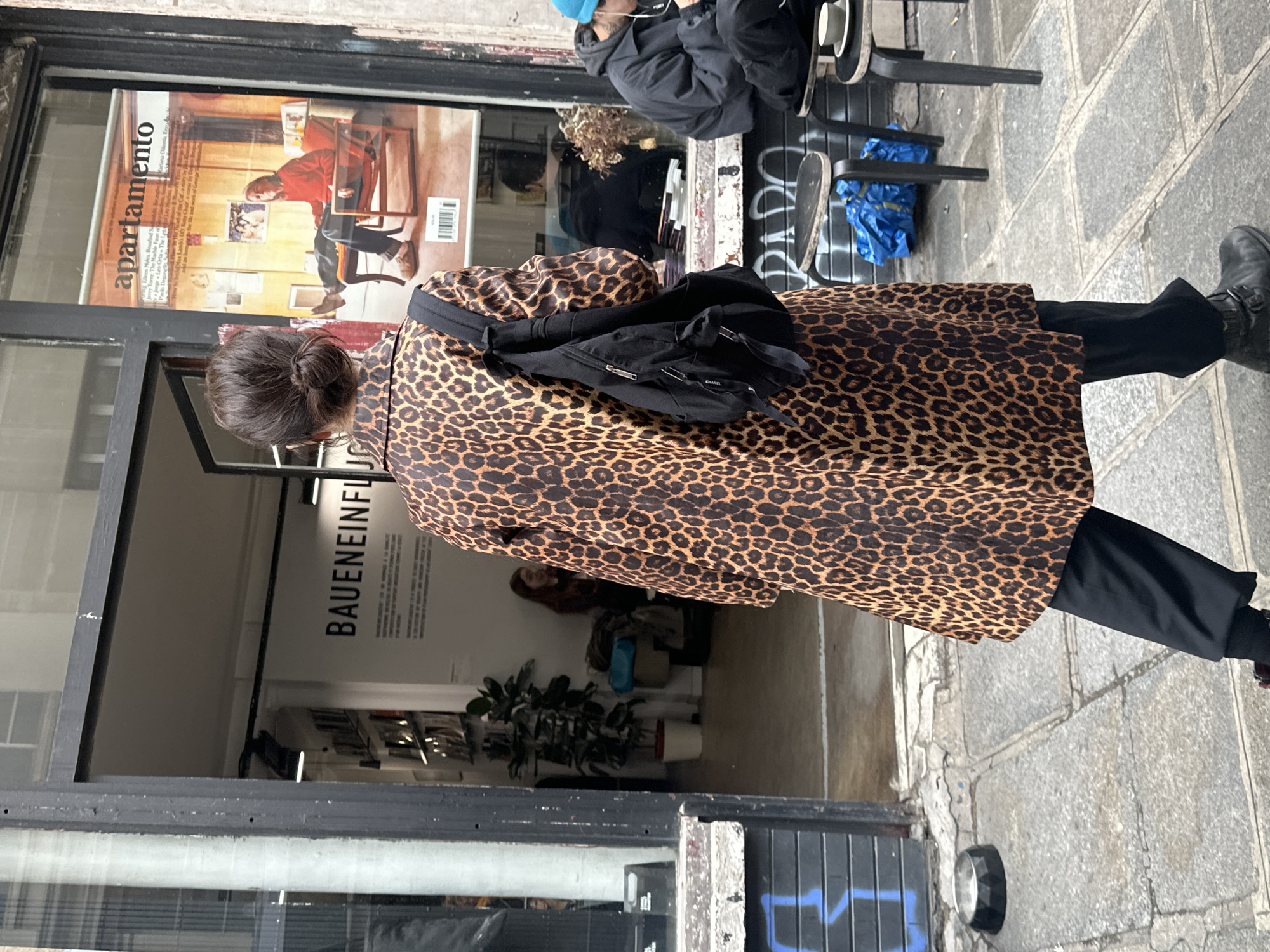 A person wearing a leopard print coat and black pants walks past a shop in Paris, France. The shop window displays a magazine cover called "Apartamento" and the sign "BAUENEINFLU". Other people are sitting at a table outside the shop. The sidewalk is grey and made of stone.