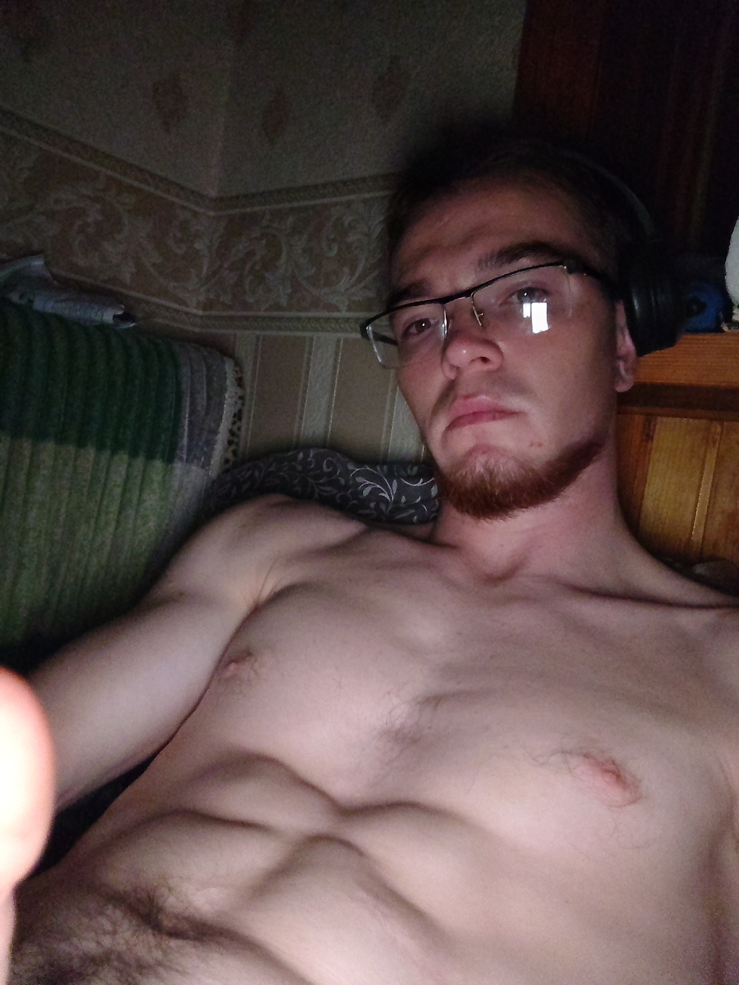 The image is a close-up, waist-up, selfie of a shirtless man with glasses and headphones on, lying on a bed. The man has a short beard and visible abdominal muscles. The background shows a patterned wallpaper and a wooden headboard. The lighting is dim, creating a shadowed atmosphere.