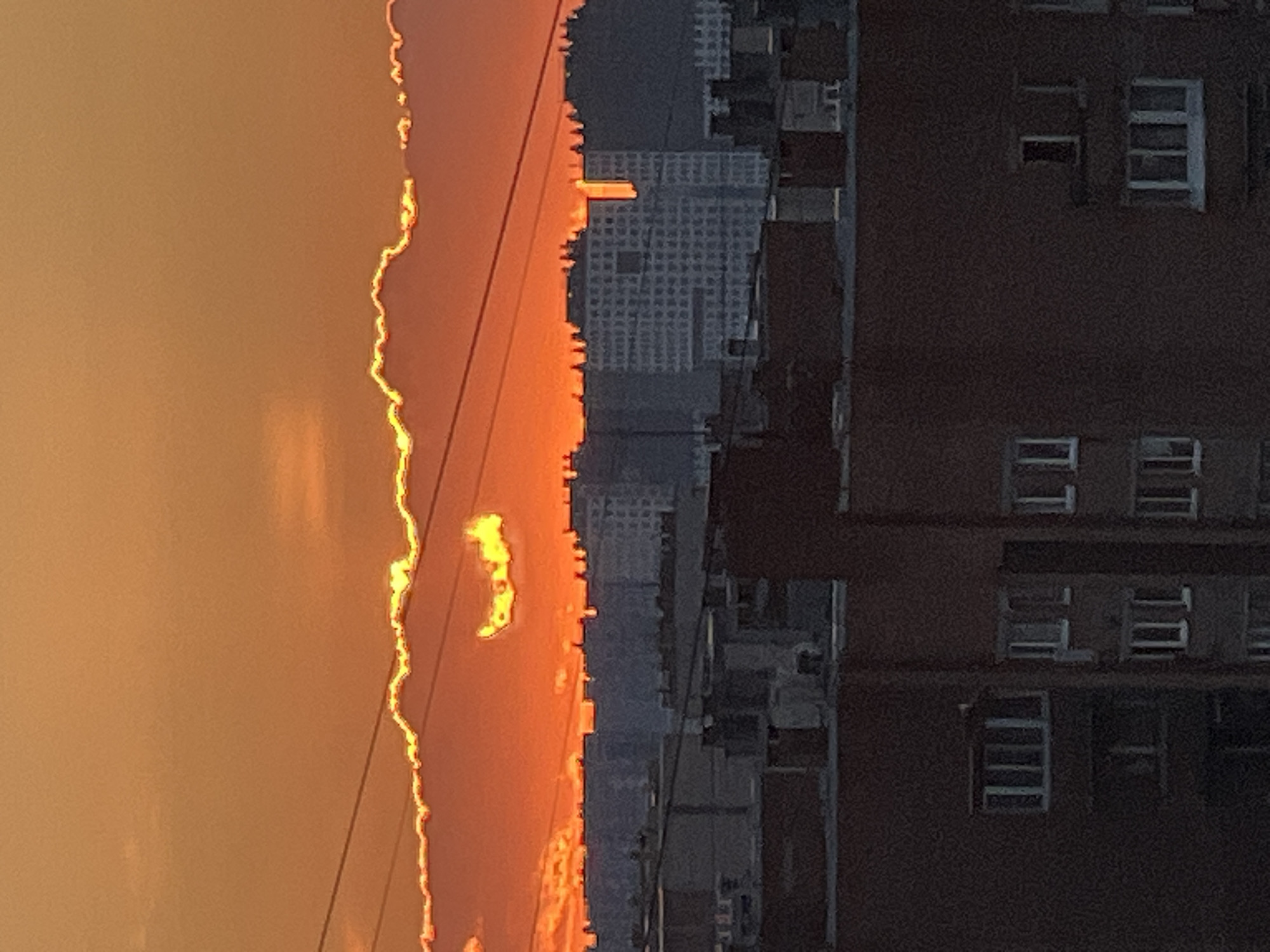 The image shows a city skyline at sunset, with the sun peeking through the clouds and casting an orange glow. In the foreground, there are several brick buildings with visible windows. Behind them, a line of taller, more modern buildings can be seen. The sky is a vibrant orange and yellow, with darker clouds obscuring some of the light. The scene appears to be a typical urban landscape, with a focus on the interplay of natural light and man-made structures. The location is Ladozhskoe Ozero, Russia, which is a city on the eastern shore of Lake Ladoga.
