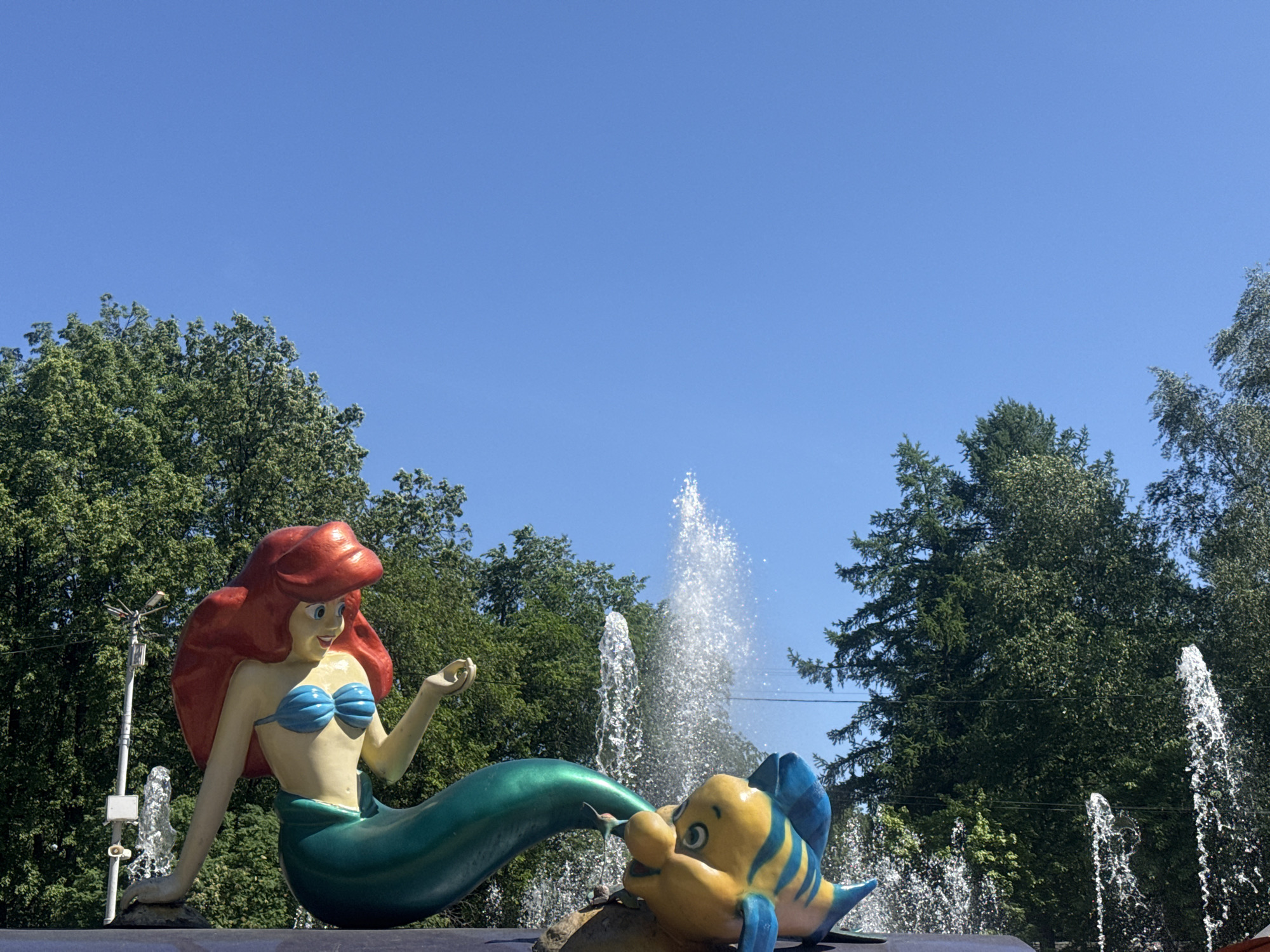 The image depicts a statue of Ariel from "The Little Mermaid" alongside Flounder, located within a fountain. The scene is situated against a backdrop of trees and a bright blue sky. The fountain is actively spraying water. The location is in Yekaterinburg, Russia. Ariel is depicted with red hair, a blue seashell bra, and a green mermaid tail. Flounder is yellow with blue fins and is positioned near the tail of Ariel.